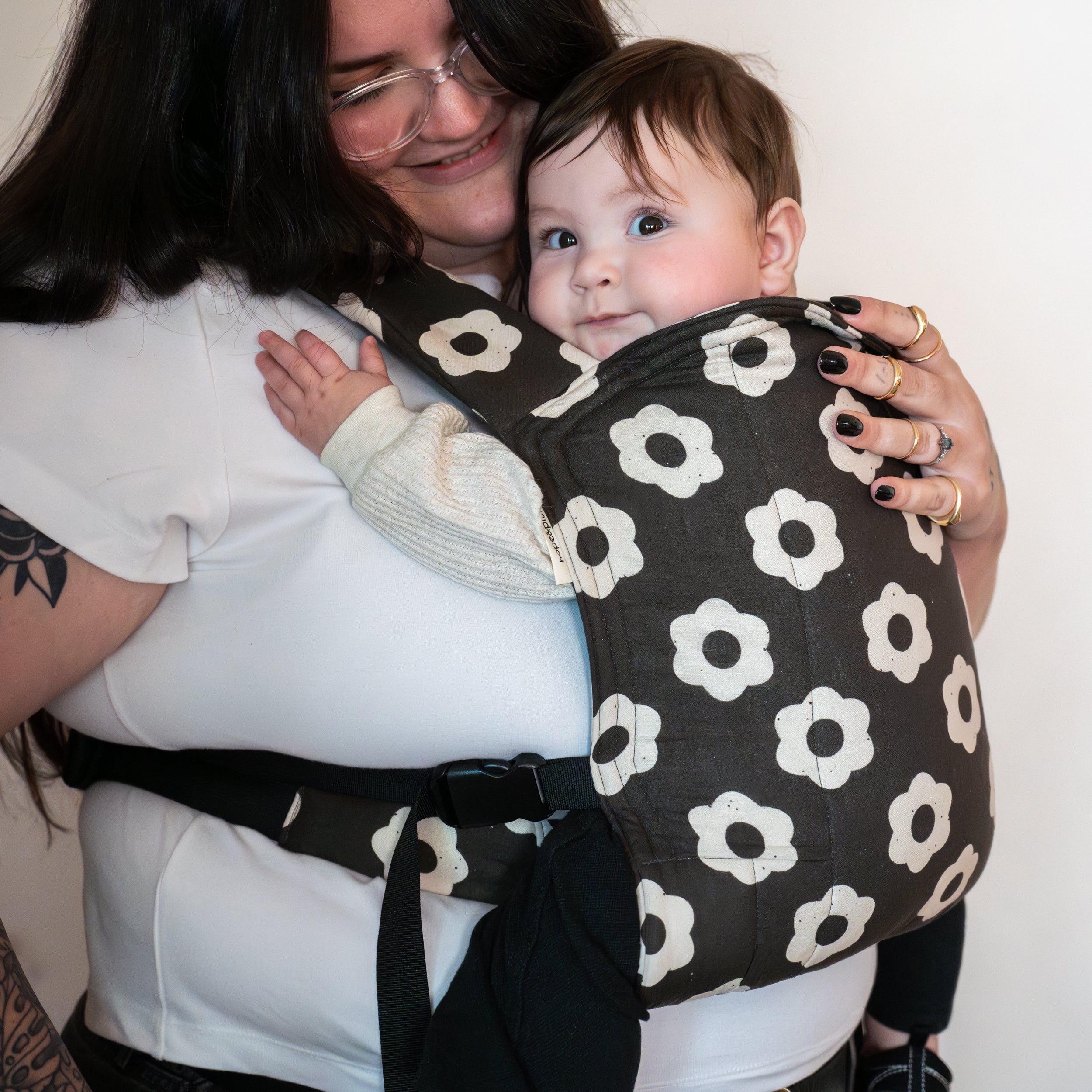 Woman holding a baby in a black and white floral-patterned buckle carrier against a plain background