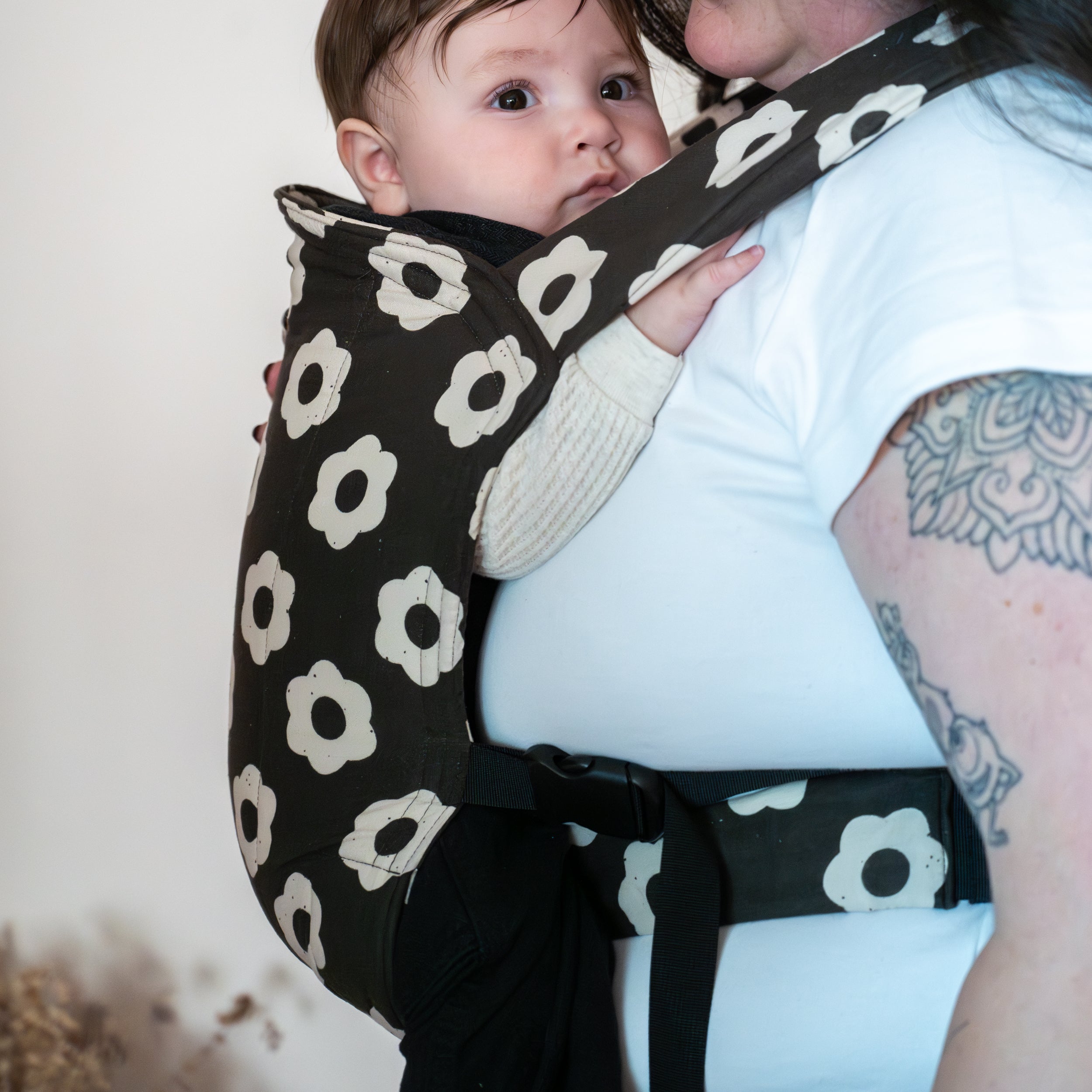Baby being carried in a black and white floral-patterned buckle baby carrier against a neutral background