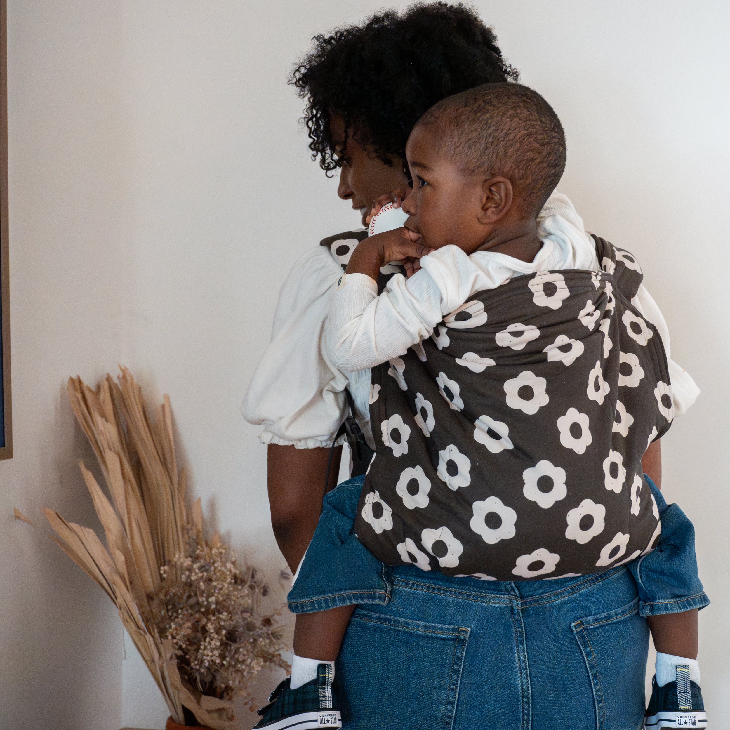 Woman holding a child in a black and white floral-patterned kid buckle carrier against a white wall.