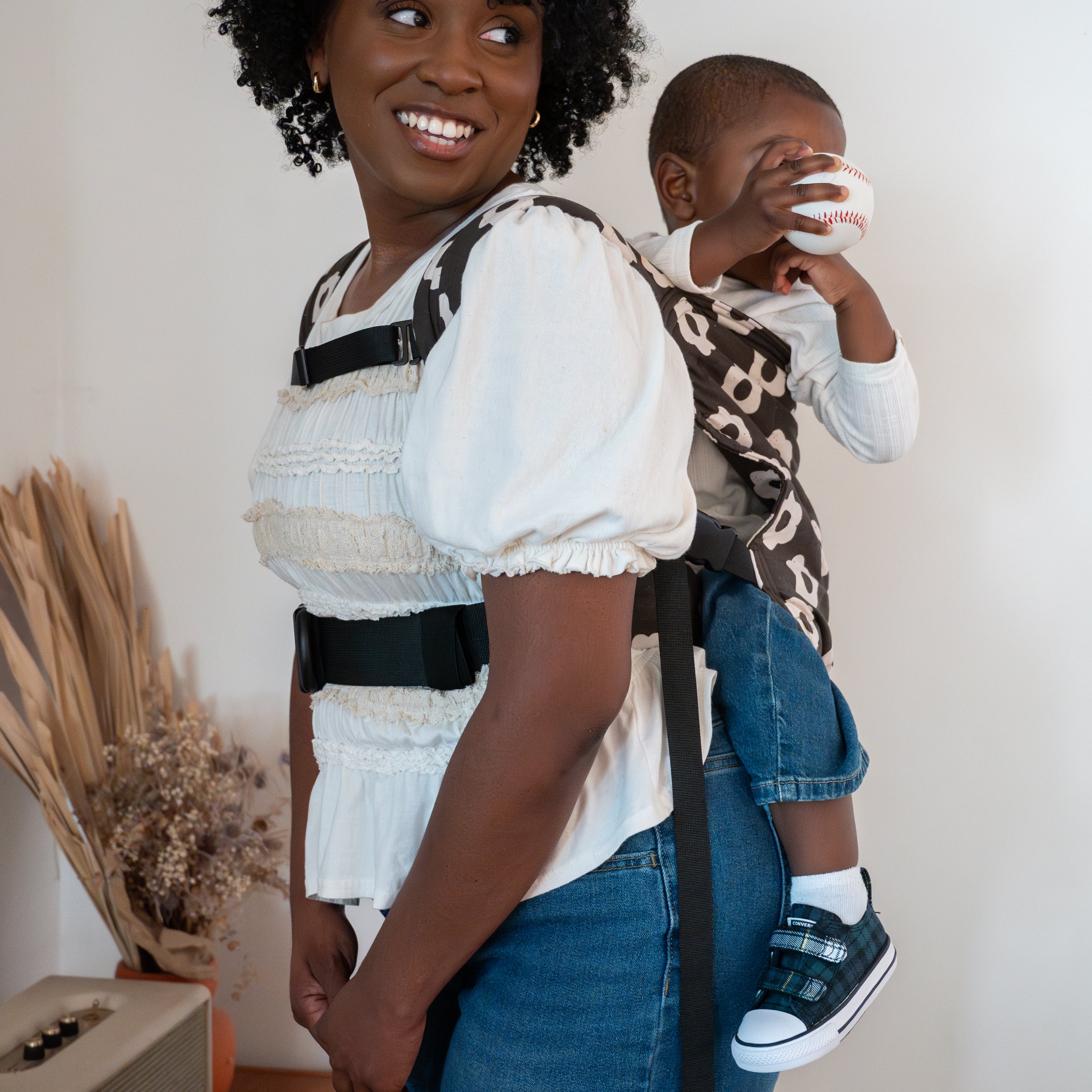 Woman carrying a child in a black and white floral buckle kid carrier against a white wall.