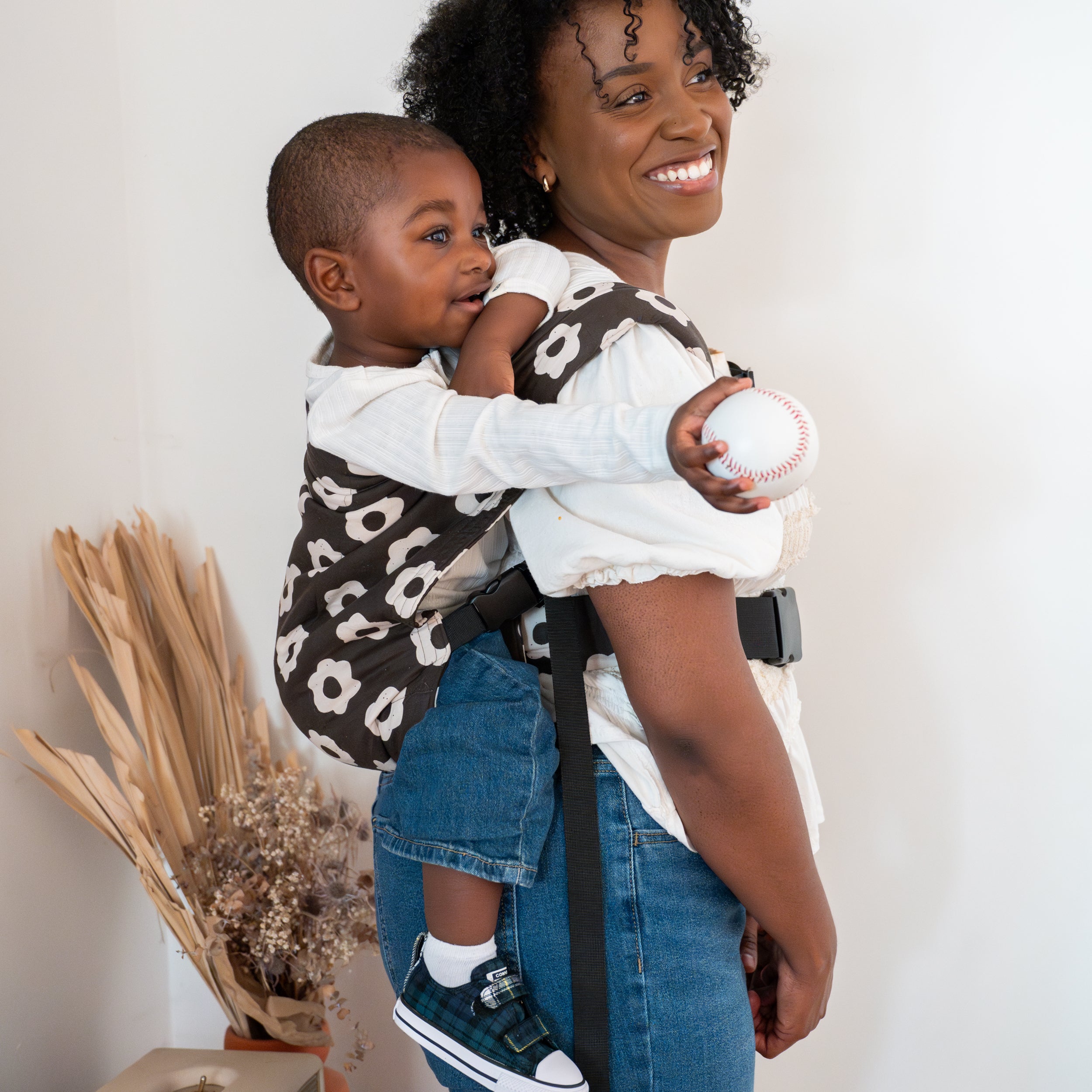 Woman holding a child in a black and white floral buckle carrier, both smiling, with a white background