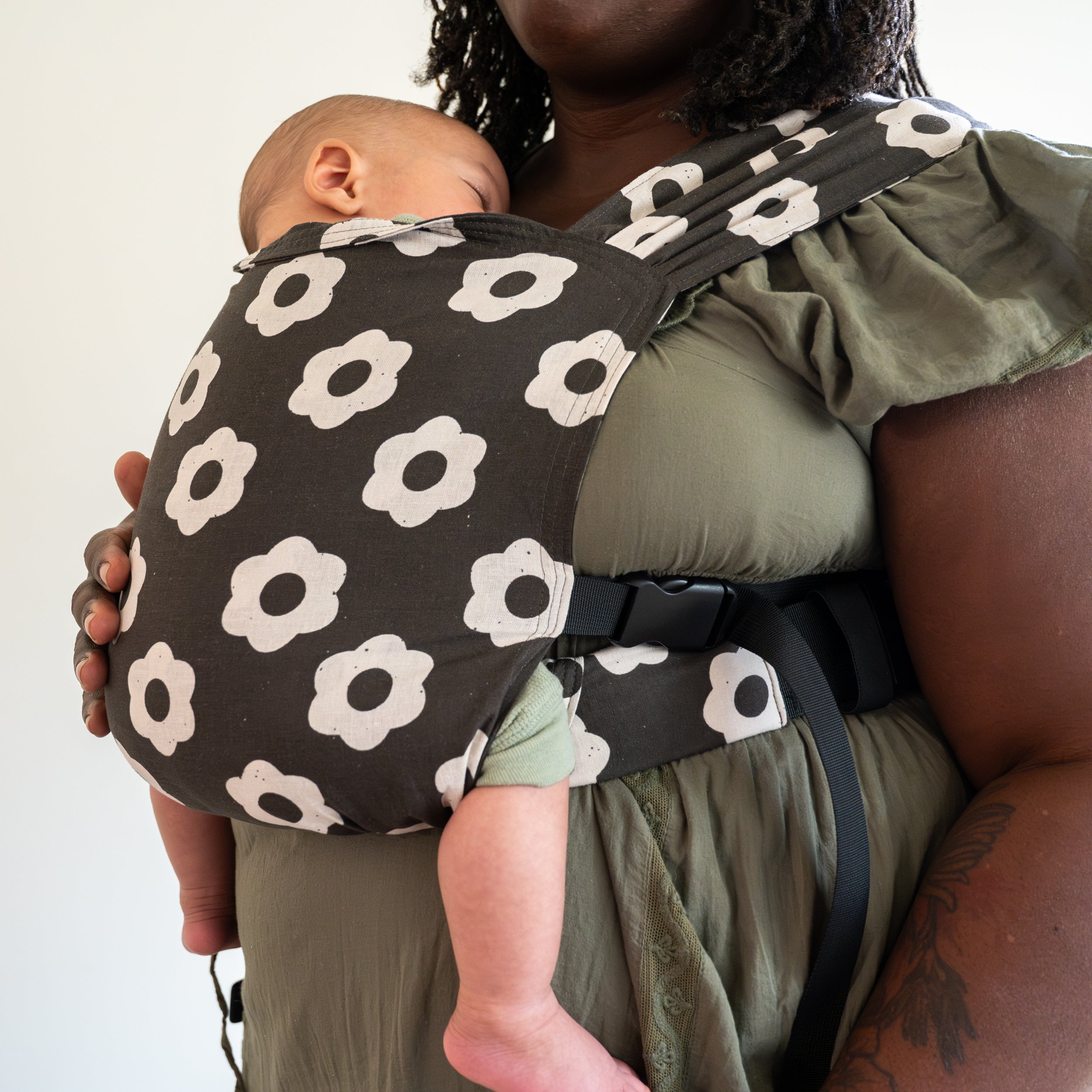 Person wearing a baby in a black and white floral-patterned newborn buckle carrier against a plain background