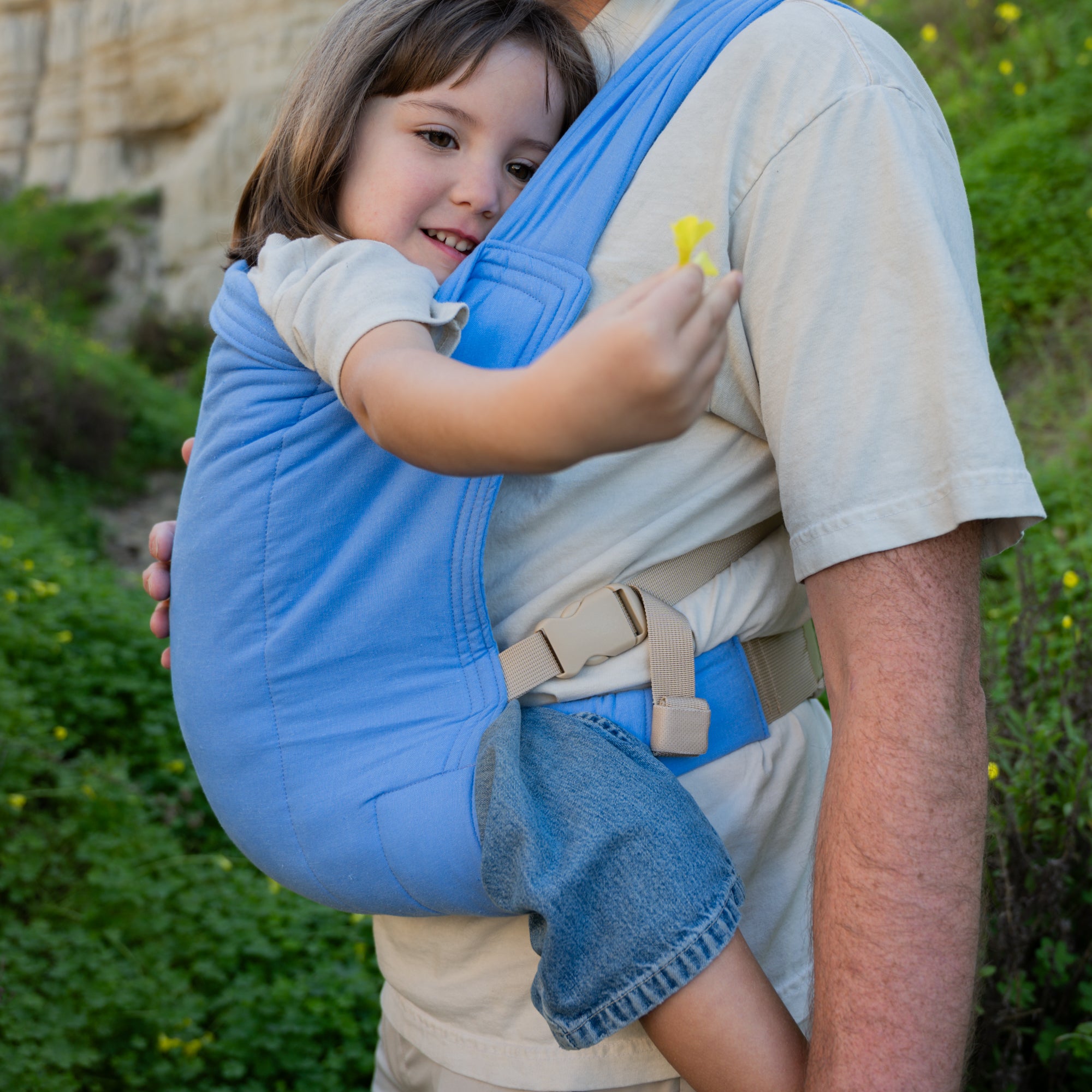 Child in a blue baby carrier held by an adult outdoors.