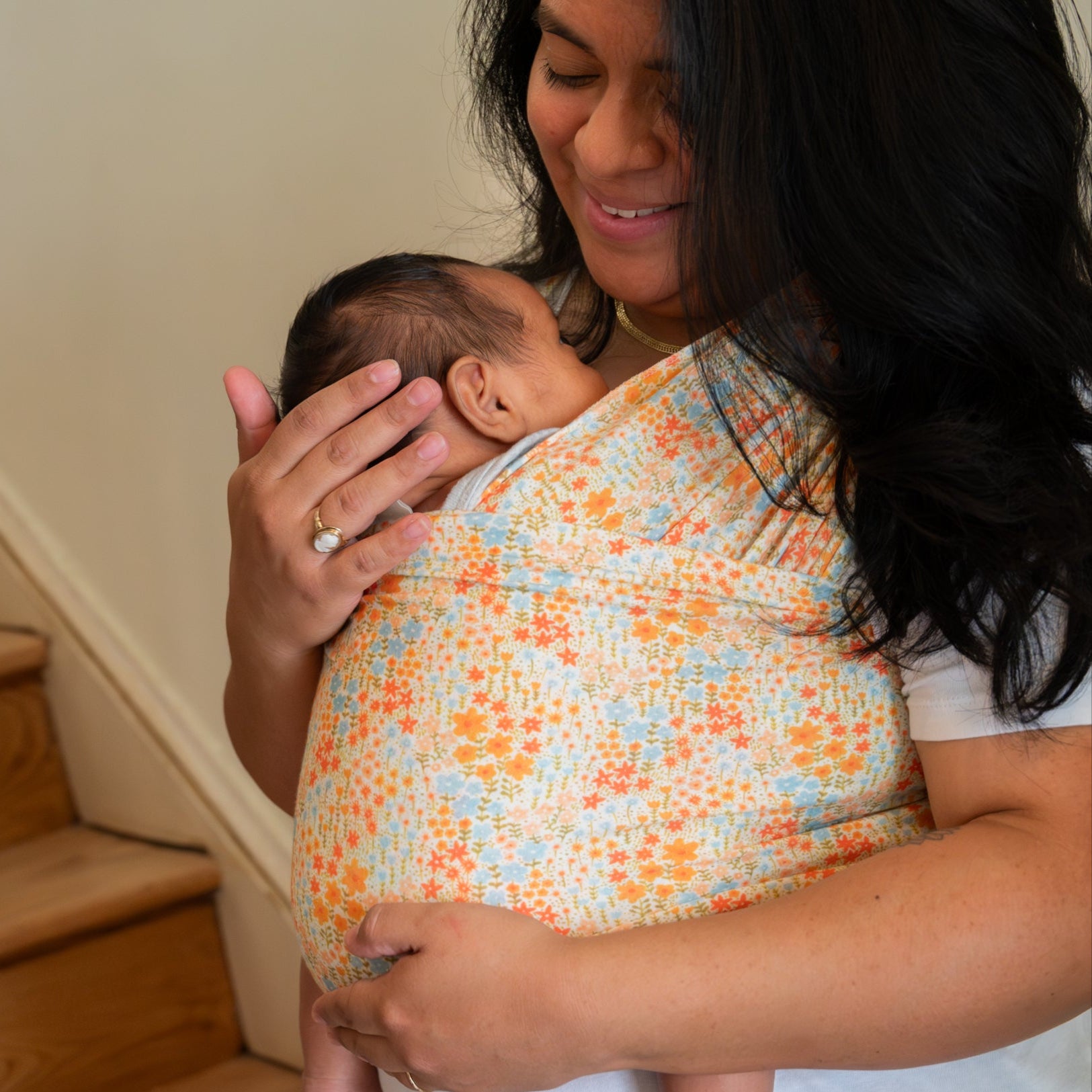 Woman holding a baby in a floral baby wrap carrier at the bottom of a staircase