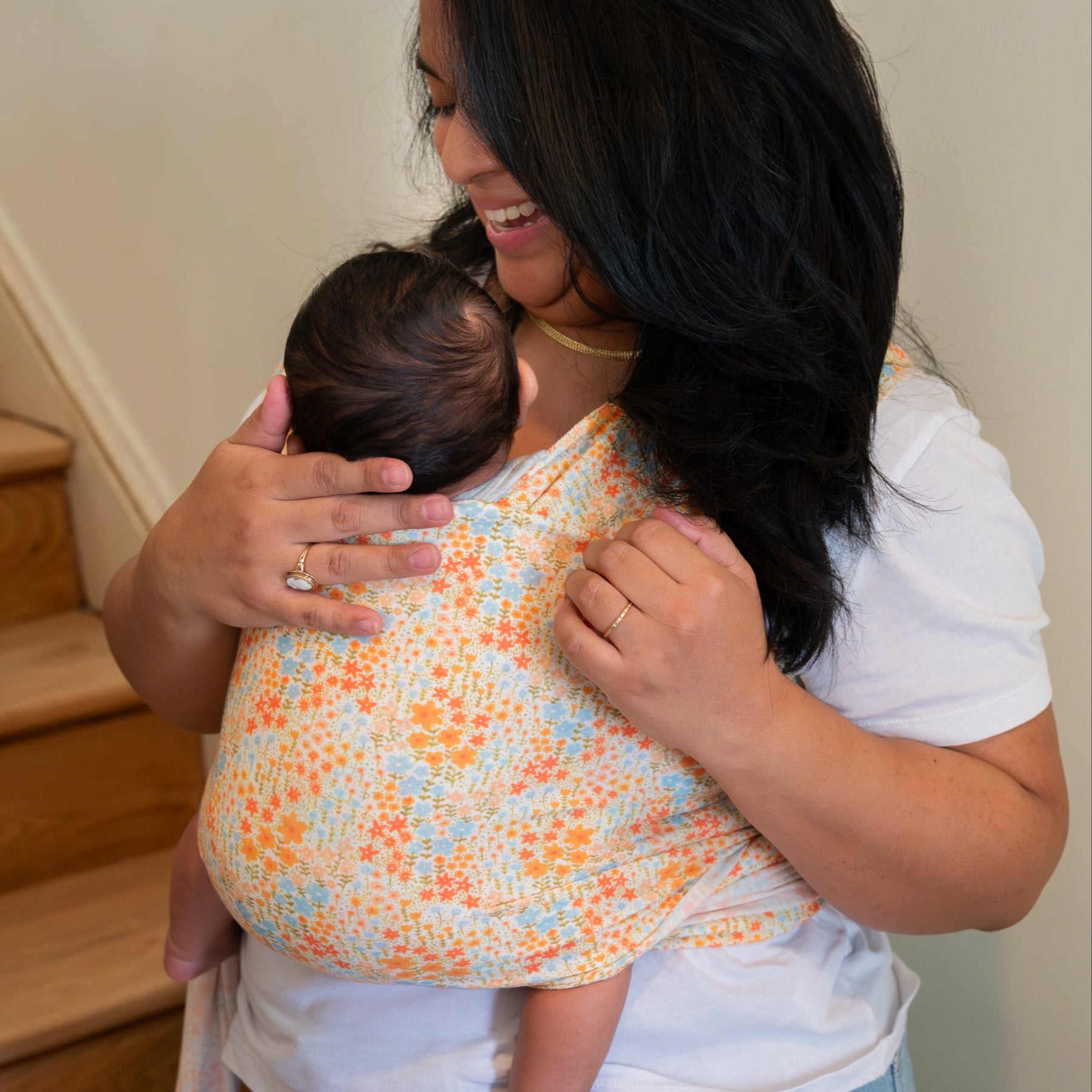 Woman holding a baby in a floral baby carrier indoors.