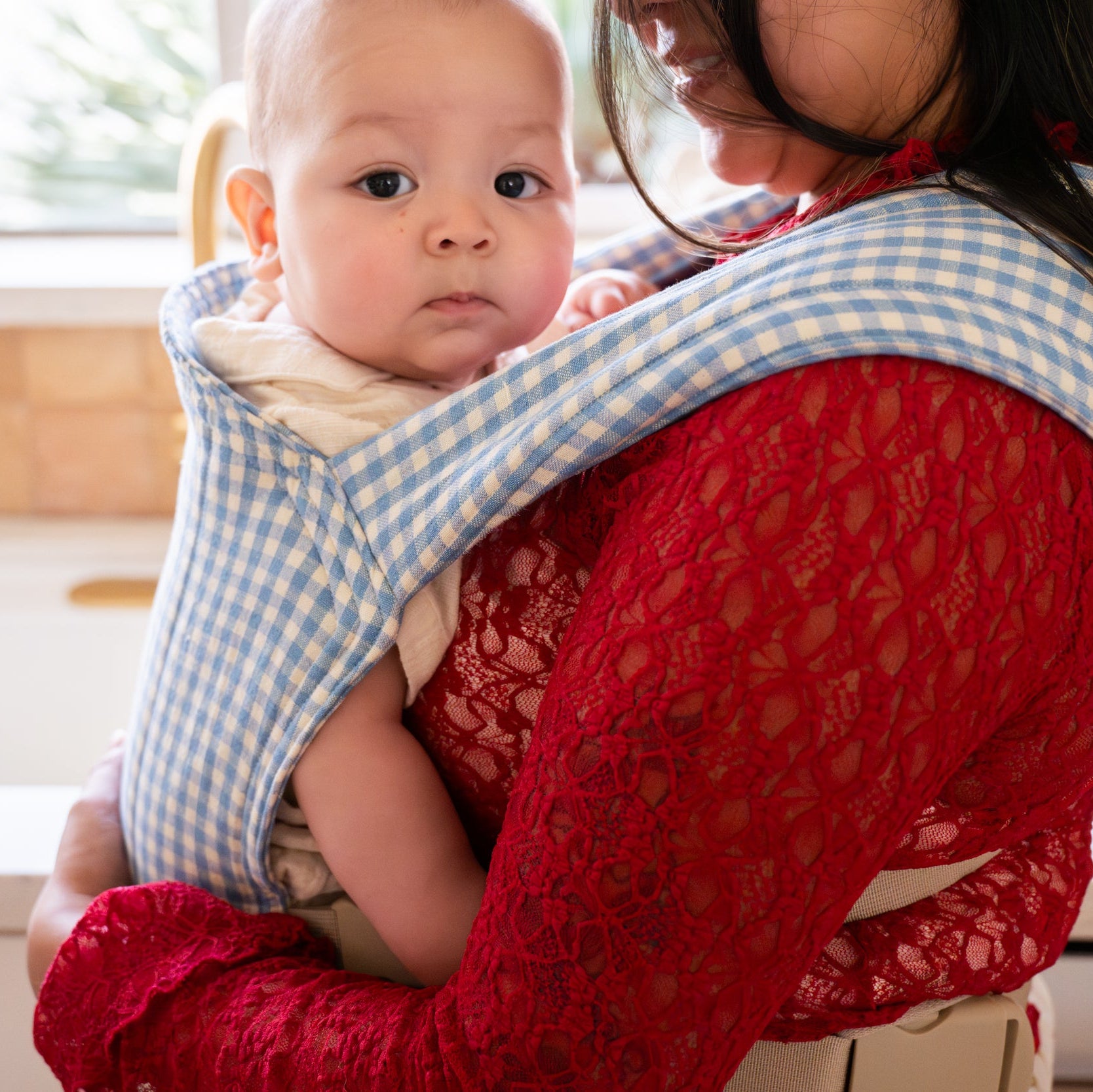 Woman holding a baby in a blue gingham buckle baby carrier indoors.