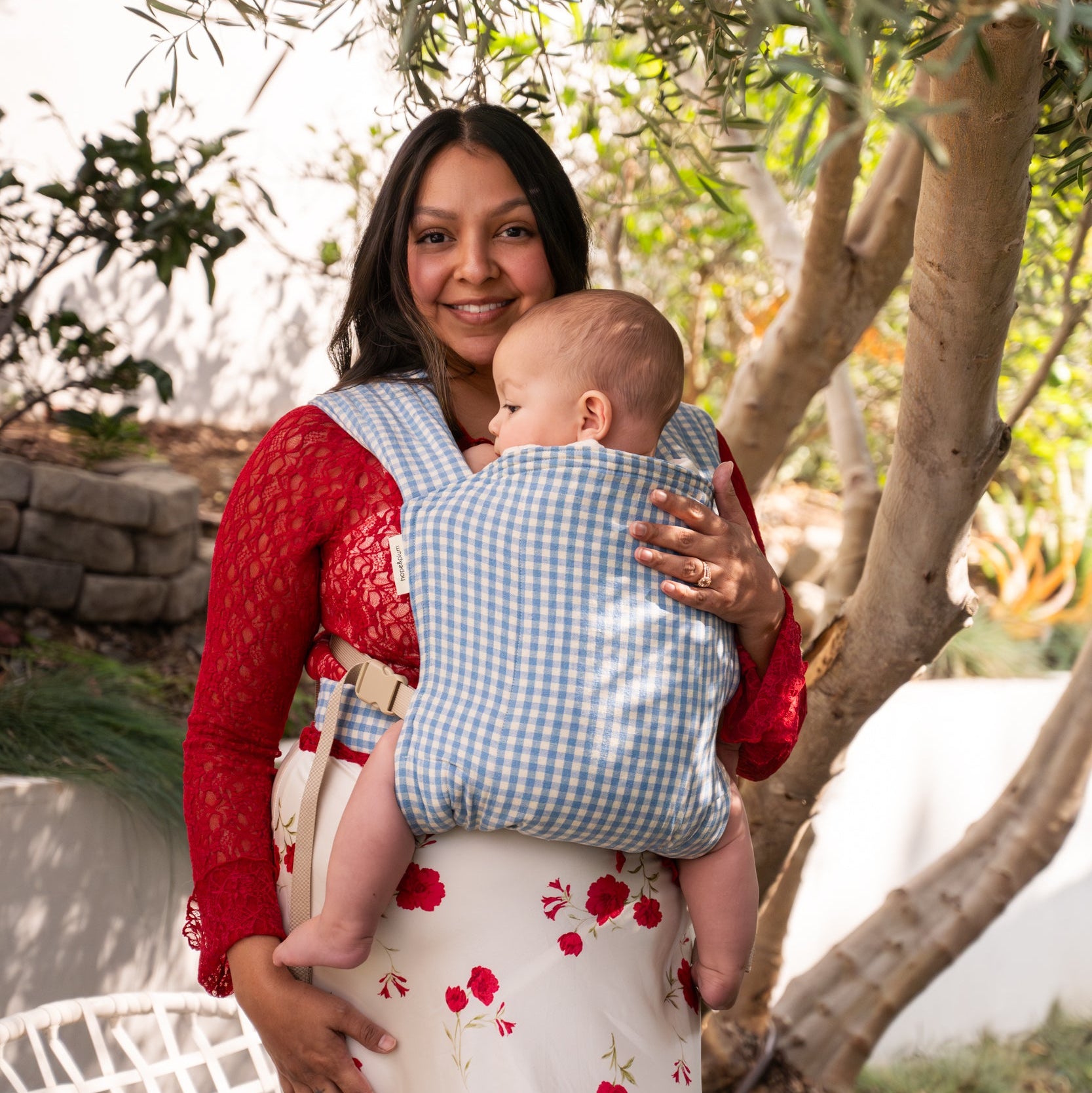 Woman holding a baby in a blue ginham buckle carrier outdoors with trees in the background