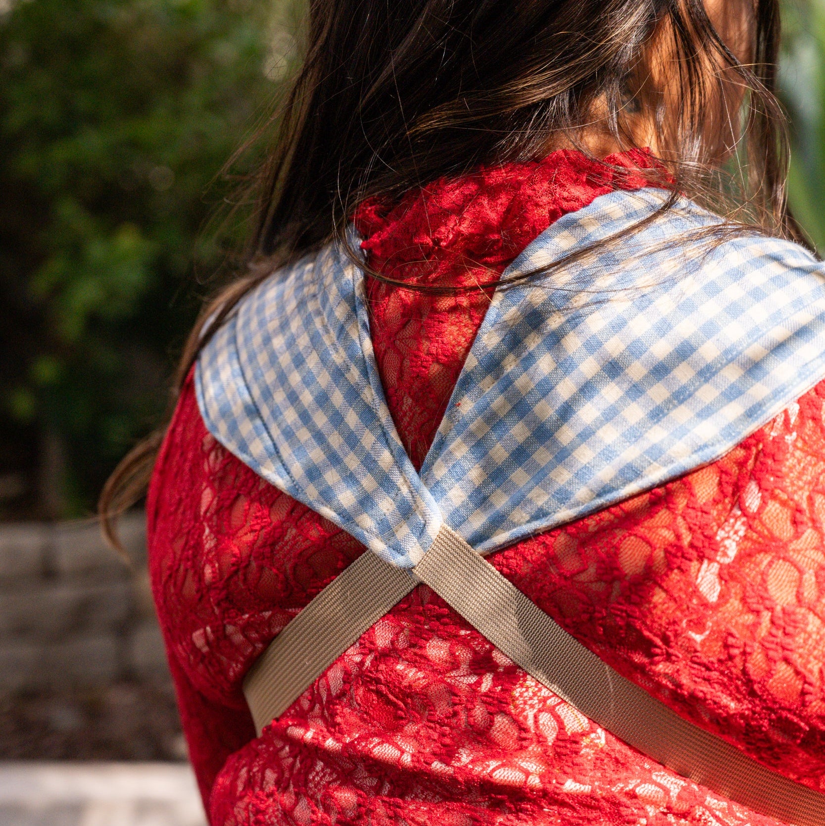 Back of a person wearing a red shirt with a blue gingham baby carrier straps across the back outdoors