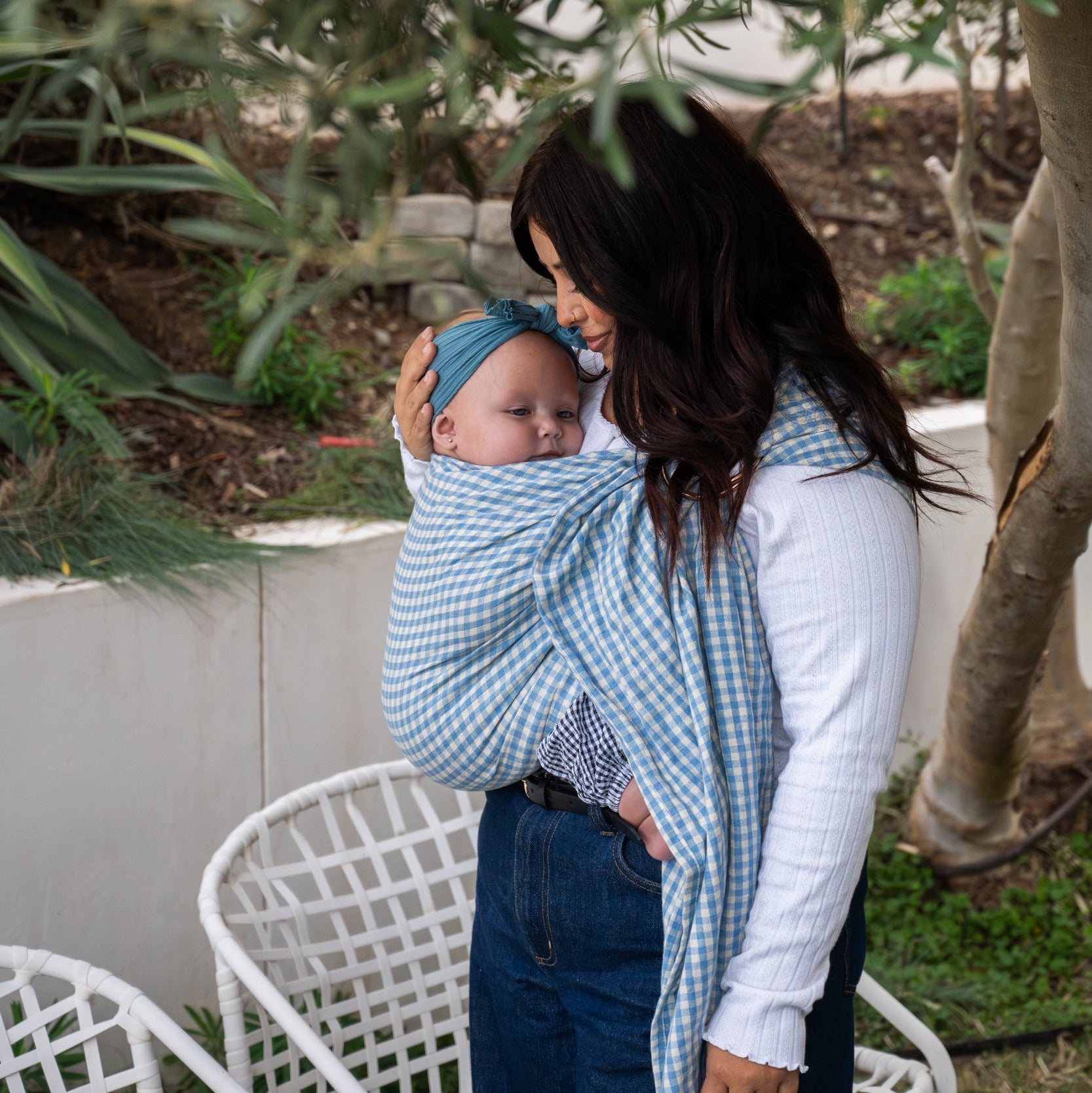 Woman holding a baby in a blue and white gingham ring sling outdoors with plants in the background