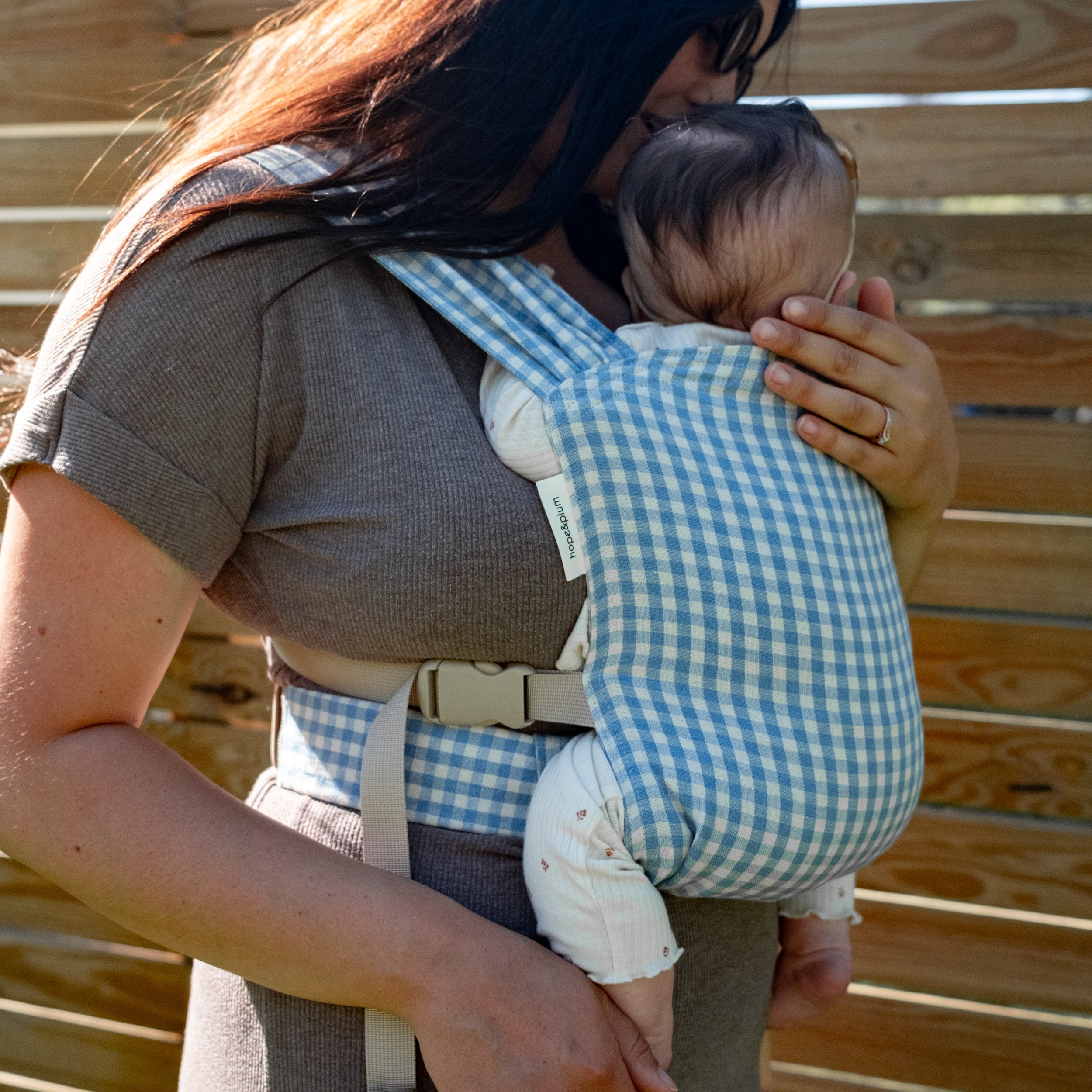 Woman carrying a baby in a blue gingham newborn buckle carrier against a wooden background