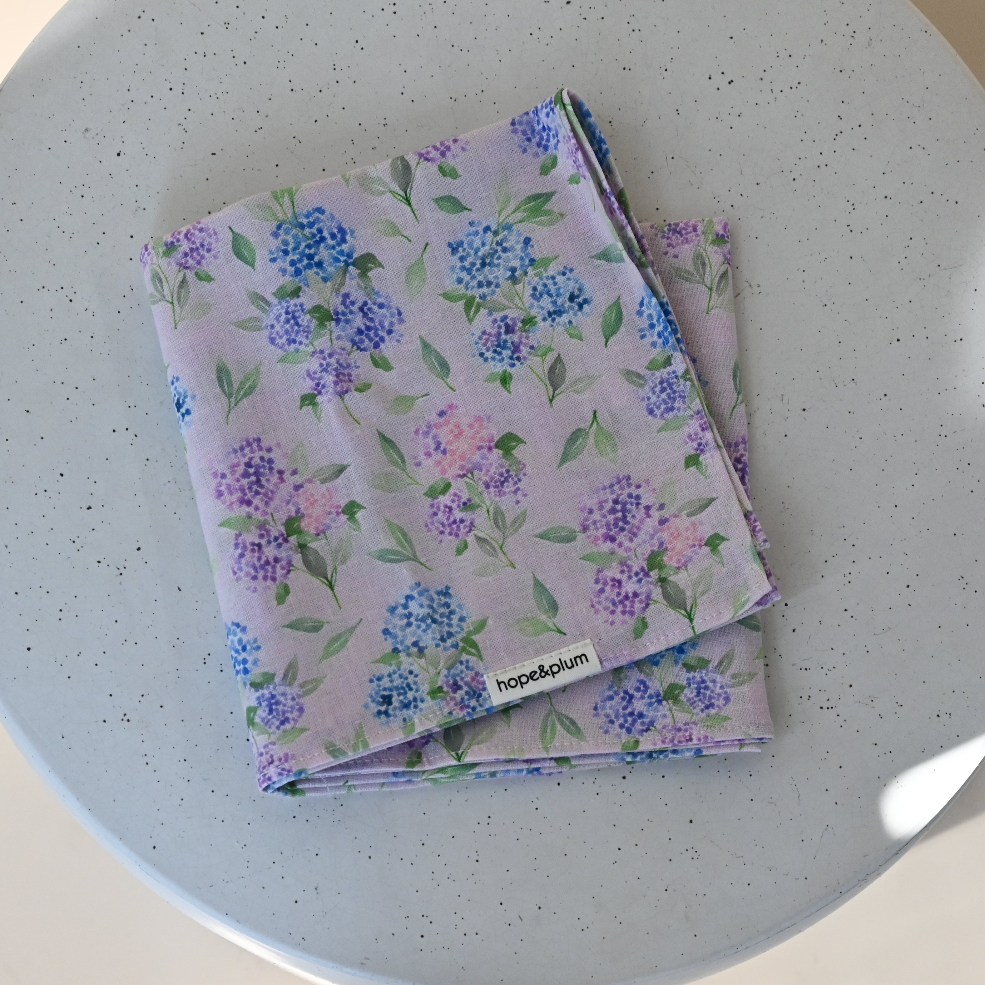Large Bandana in Blush — the folded bandana showing hydrangea clusters in blue, lavender and pink with green leaves on a soft pink background.