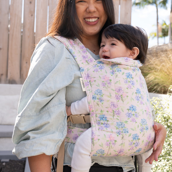Lark Baby Carrier in Blush — smiling parent front-carrying a happy baby outdoors, blue and lavender hydrangea print on a soft pink background.