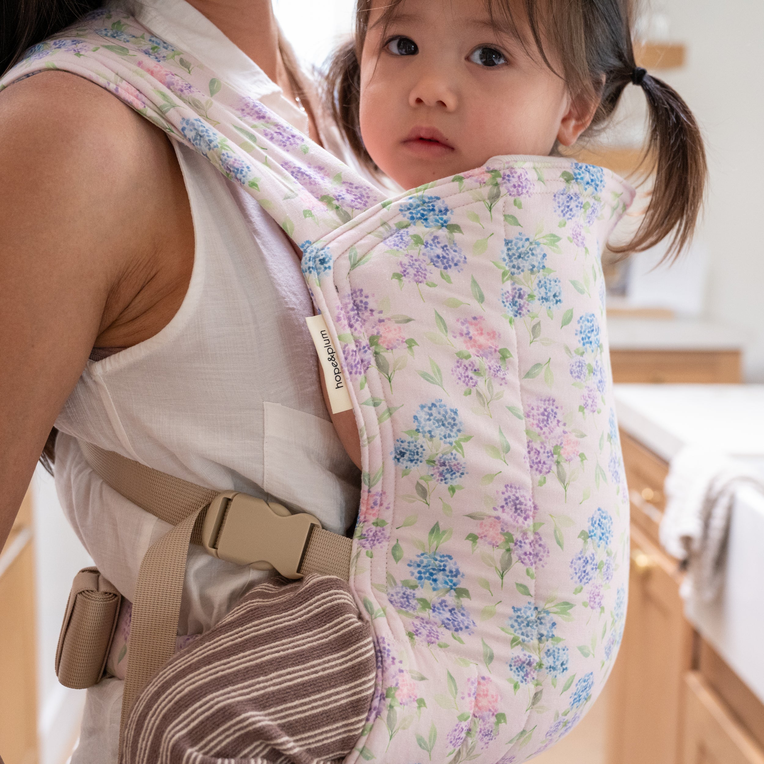 Lark Baby Carrier in Blush — toddler in pigtails worn on the front, padded hydrangea-print panel in blue and lavender on a soft pink background.