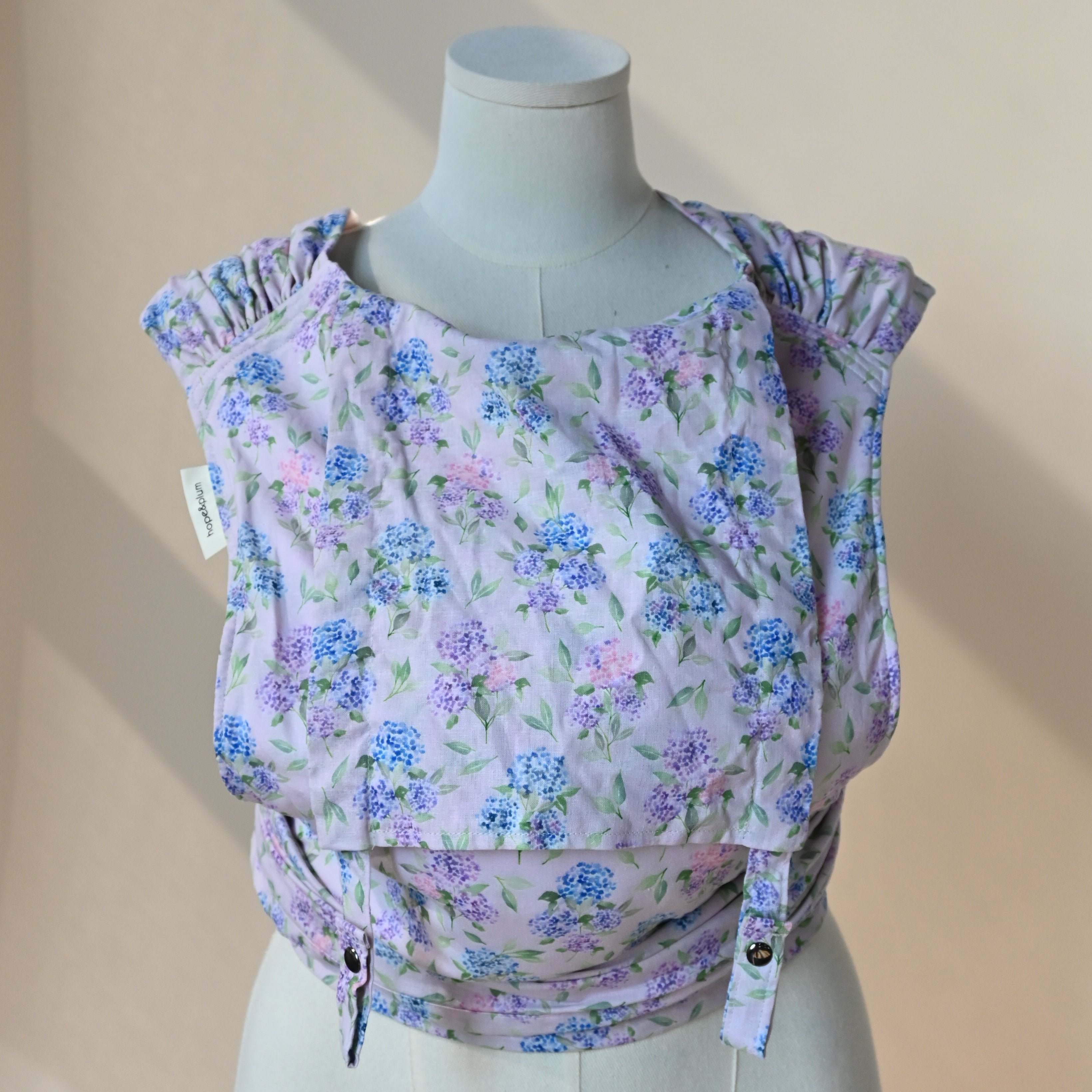 Meh Dai Baby Carrier in Blush — tie-strap carrier shown on a dress form, blue and lavender hydrangea print on a soft pink background.