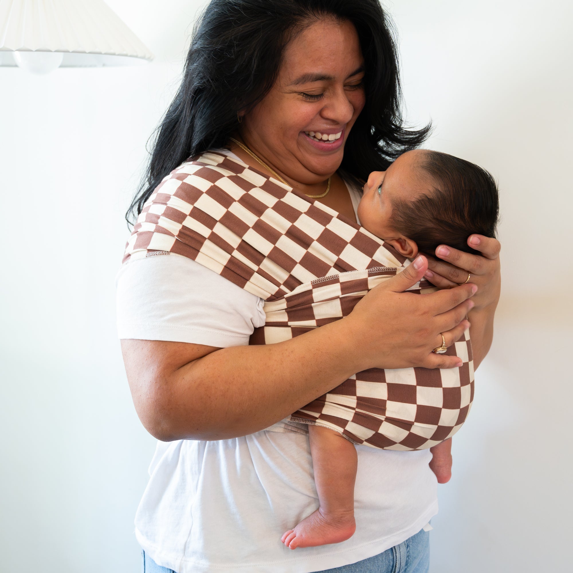 Woman holding a baby wrapped in a brown and white checkered wrap against a plain background
