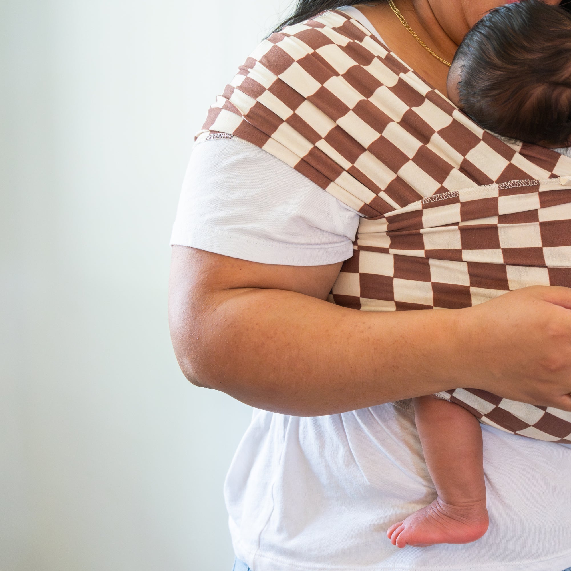 Person holding a baby in a brown and white checkered sling against a plain background