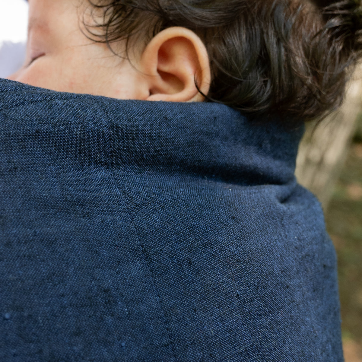 Close view of the Caspian Lark Baby Carrier's panel edge beside a baby's head, showing the fine weave of the indigo and black chambray.