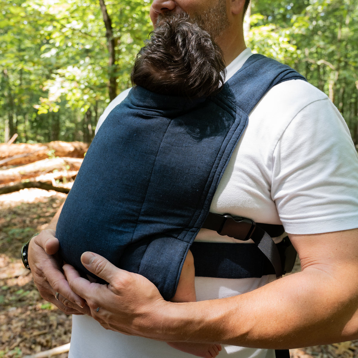 A wearer carries a baby in a front carry in the Caspian Lark Baby Carrier, the padded waistband and buckle visible below the indigo panel.