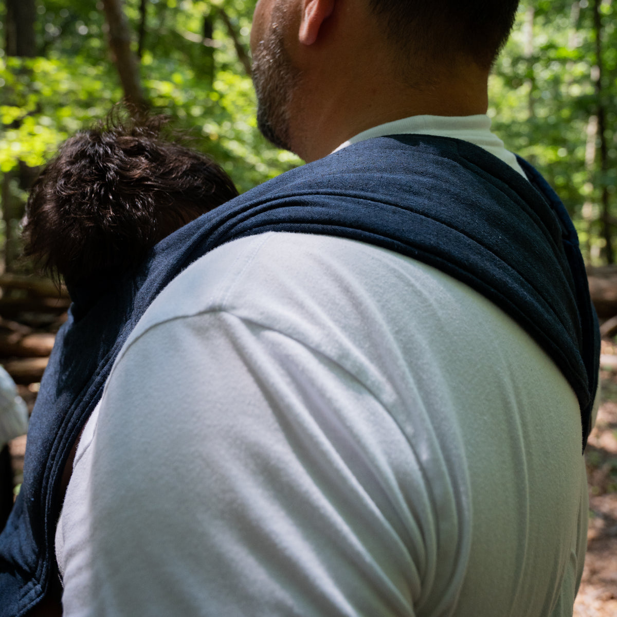 A baby's head rests against the wearer's shoulder in the Caspian Lark Baby Carrier, the indigo chambray strap drawn across the back.