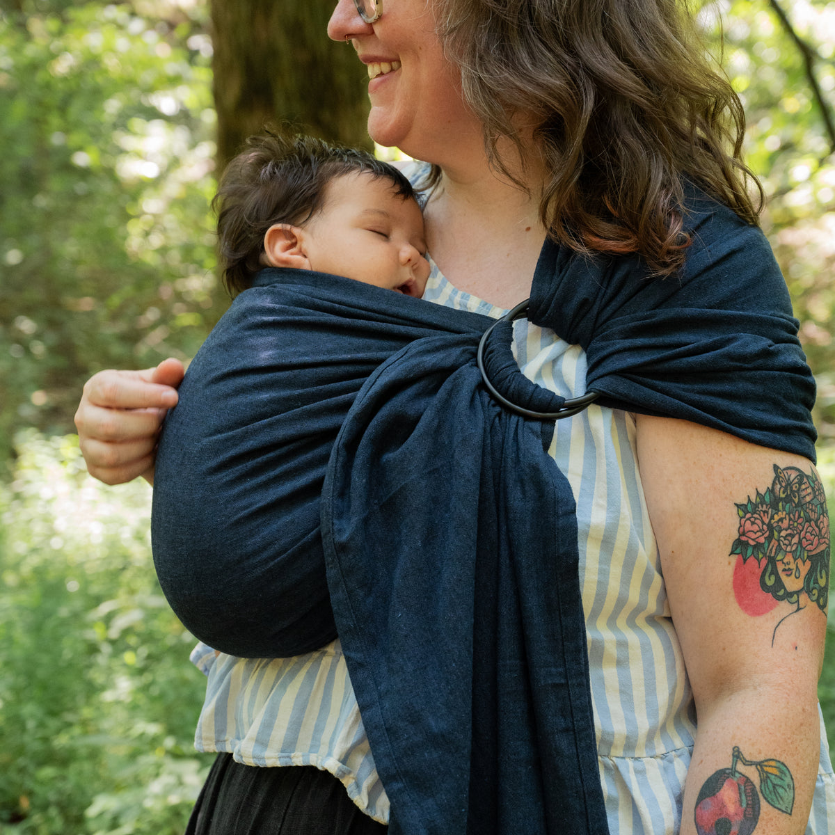 Caspian Ring Sling holding a sleeping baby against the wearer's chest, its woven chambray a deep indigo navy like dark denim, gathered through black rings.