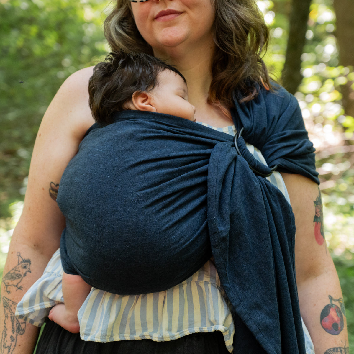 A baby sleeps in the Caspian Ring Sling, the deep indigo chambray gathered through the rings at the wearer's shoulder.