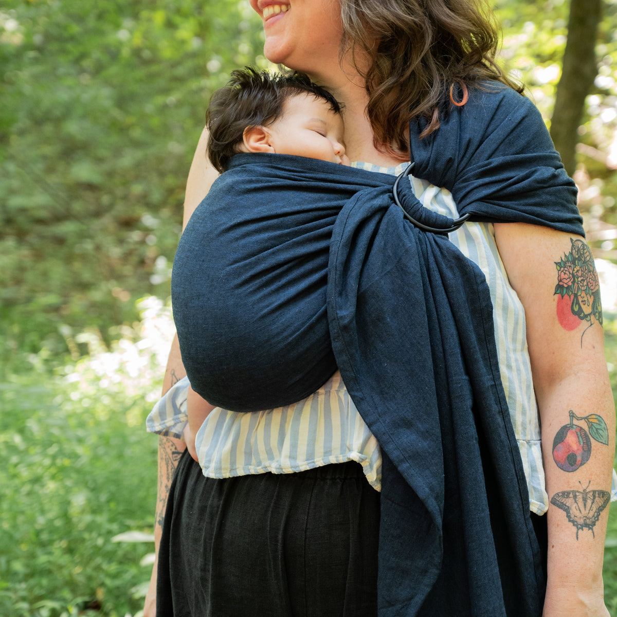 A baby sleeps against the wearer's chest in the Caspian Ring Sling, the dark indigo chambray tail falling loose below the black rings.