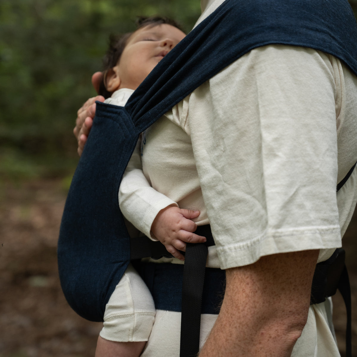 Side view of a sleeping baby in the Caspian Sprout Newborn Carrier, the indigo chambray panel and black strap across the wearer's body.