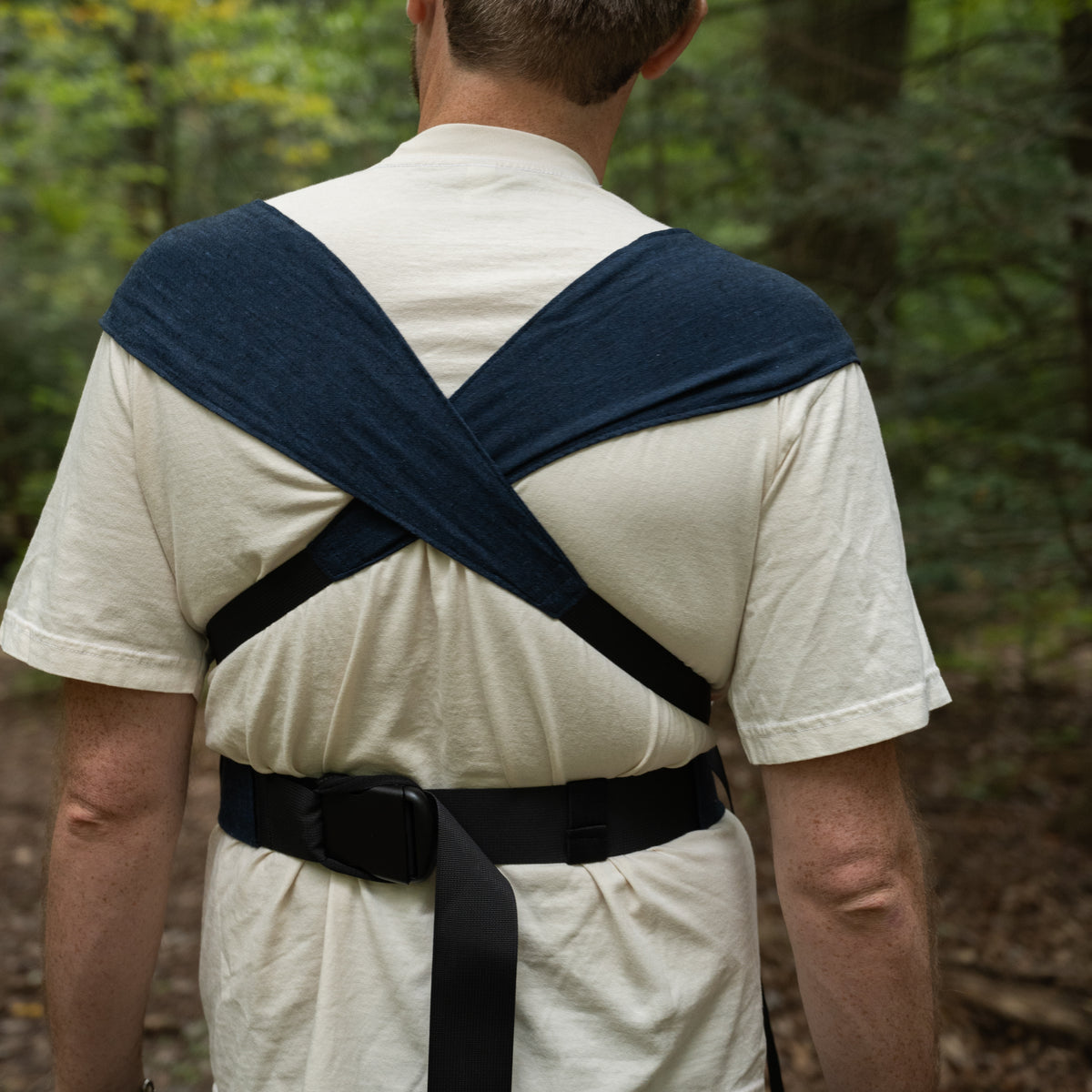 Back view of the Caspian Sprout Newborn Carrier, its indigo chambray panels crossed over the shoulders above a black buckled waistband.