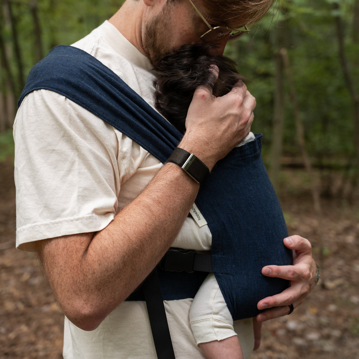 A wearer leans down to kiss a baby carried in the Caspian Sprout Newborn Carrier, a hand cupping the baby's head above the indigo panel.
