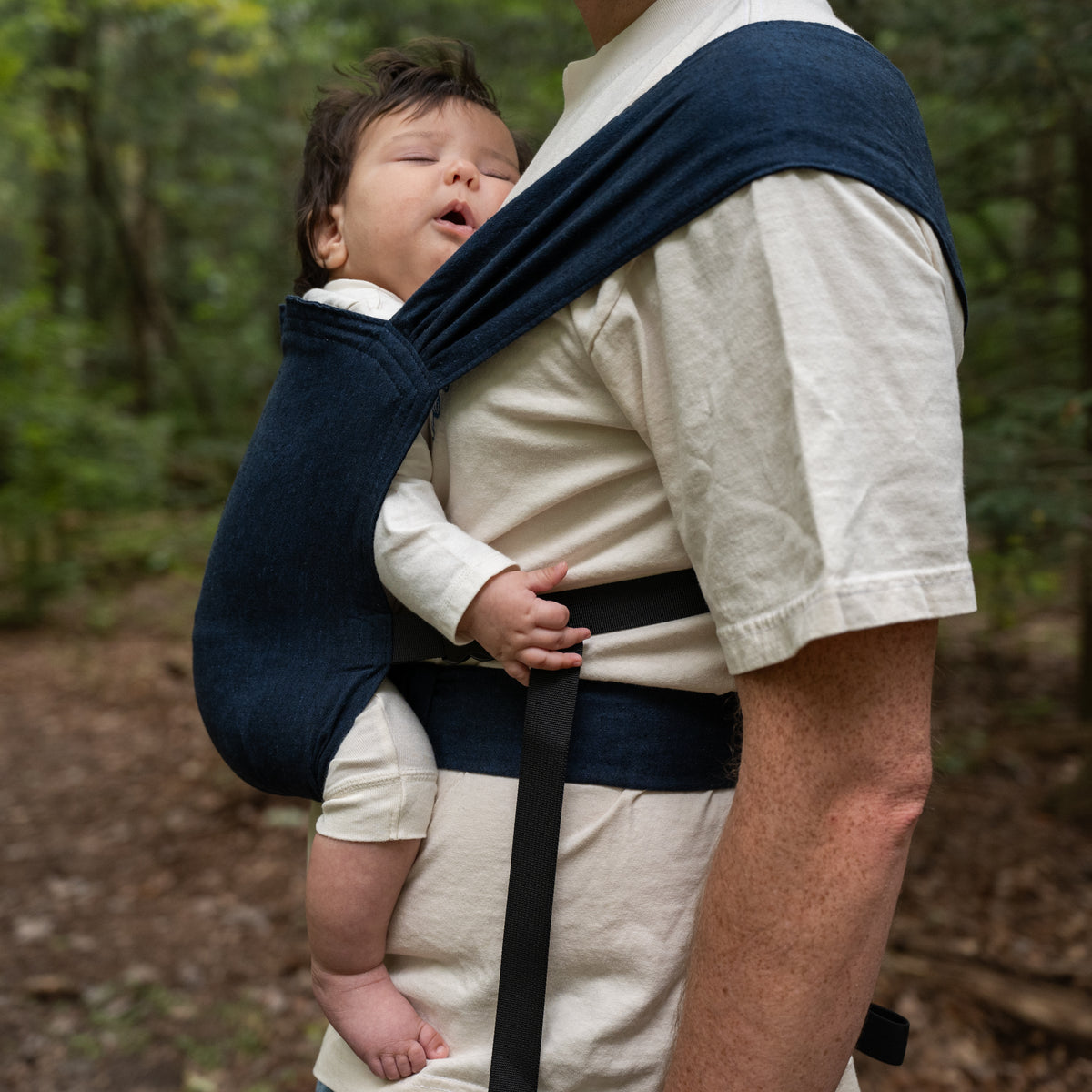 A baby sleeps in the Caspian Sprout Newborn Carrier, the indigo chambray panel supporting them with the strap drawn over the wearer's shoulder.