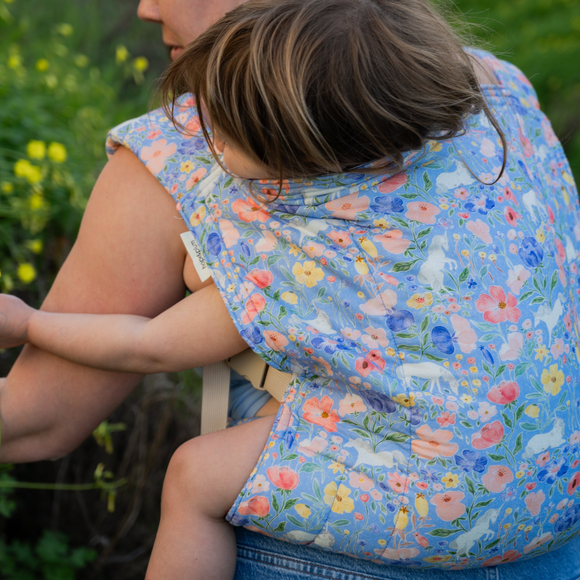 Person wearing a child on their back in a floral unicorn baby carrier with a natural outdoor background