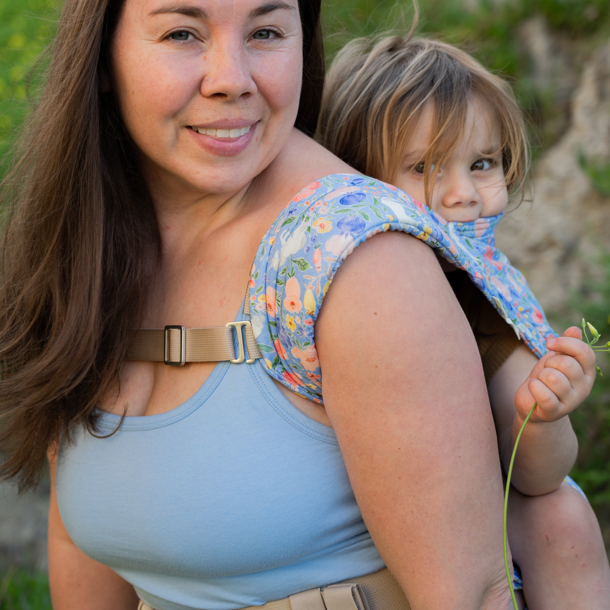 Woman carrying a child on her back with a floral unicorn baby carrier, outdoors.
