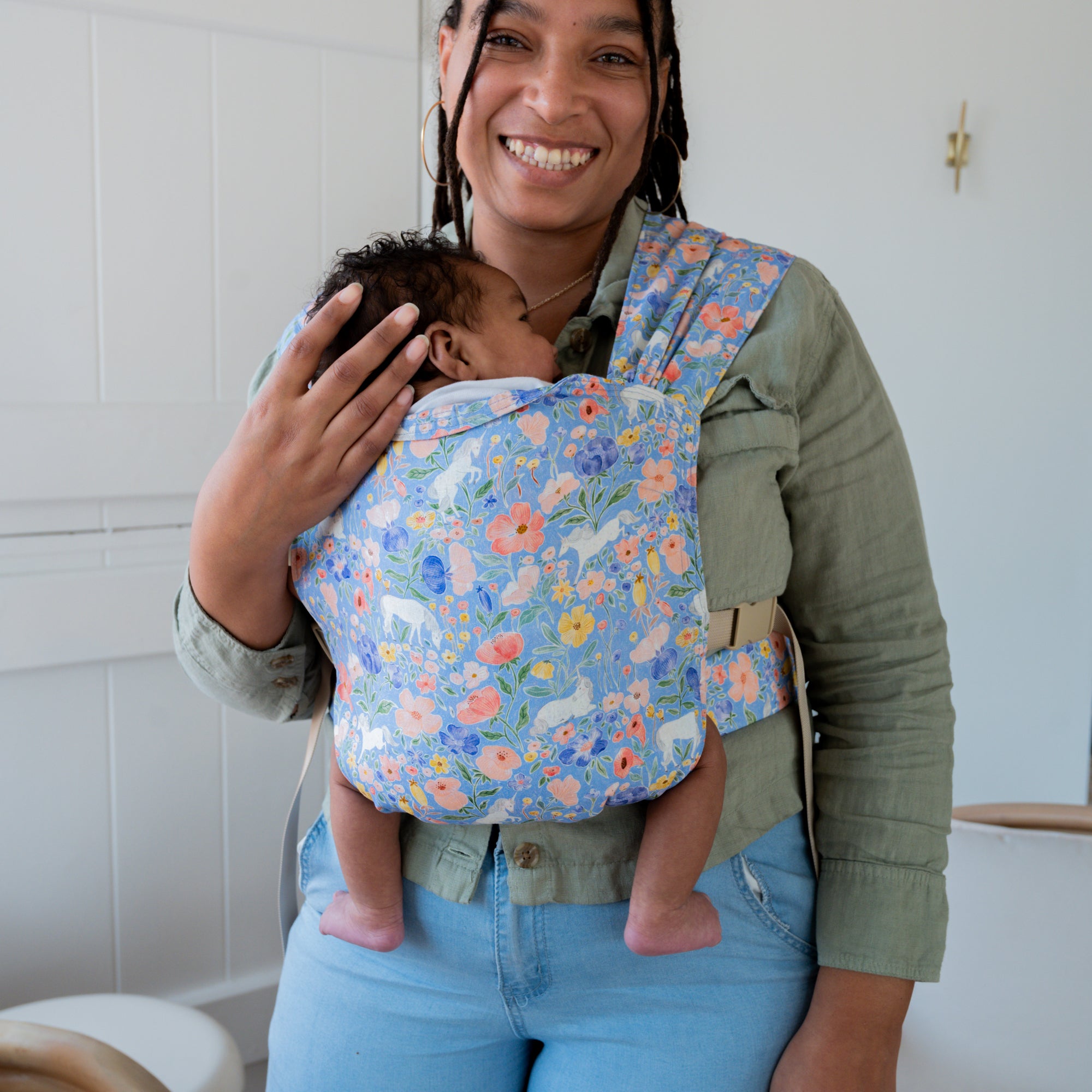 Woman holding a baby in a floral unicorn baby carrier against a white wall.