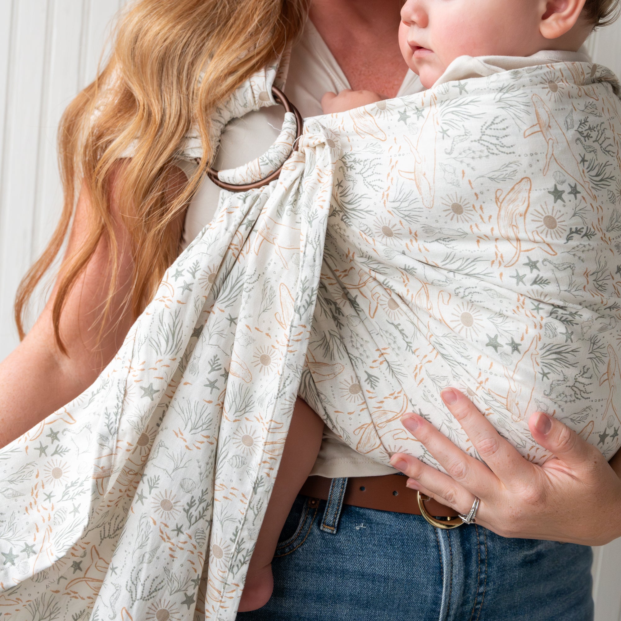 Woman holding a baby in an ocean print baby ring sling against a white wall.