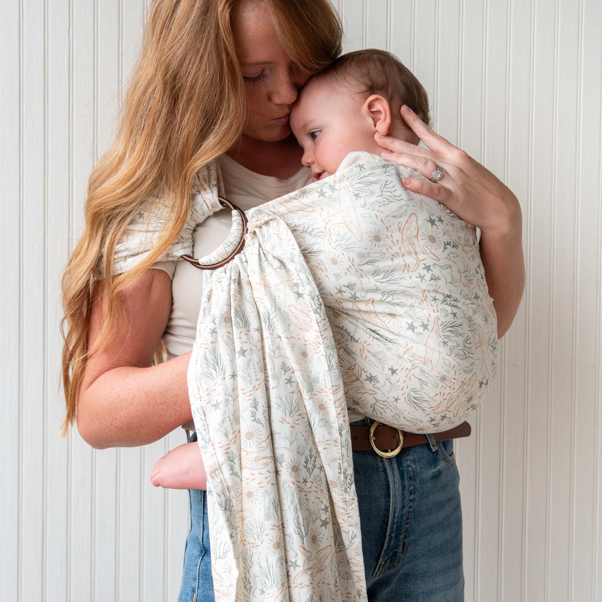 Woman holding a baby in an ocean print baby ring sling against a white wall.