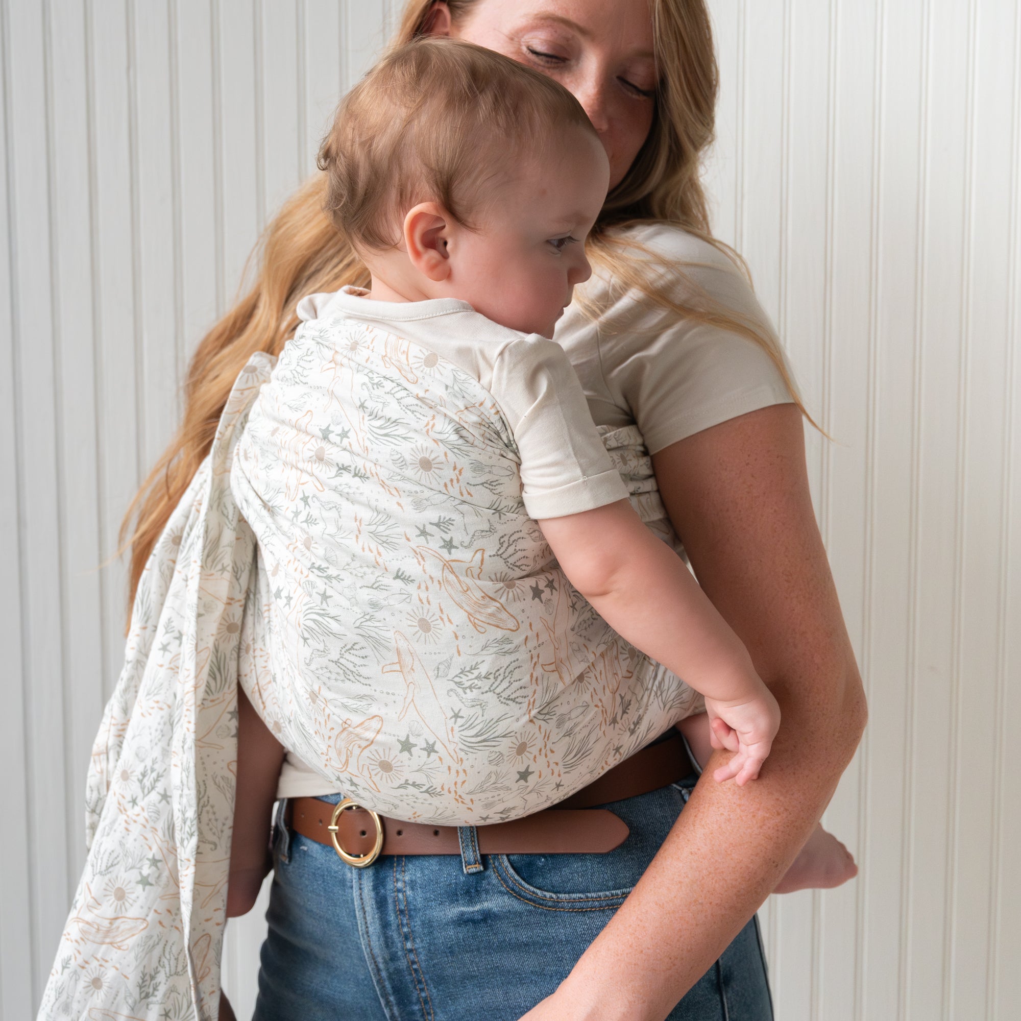 Woman holding a baby in an ocean print baby ring sling against a white wall.