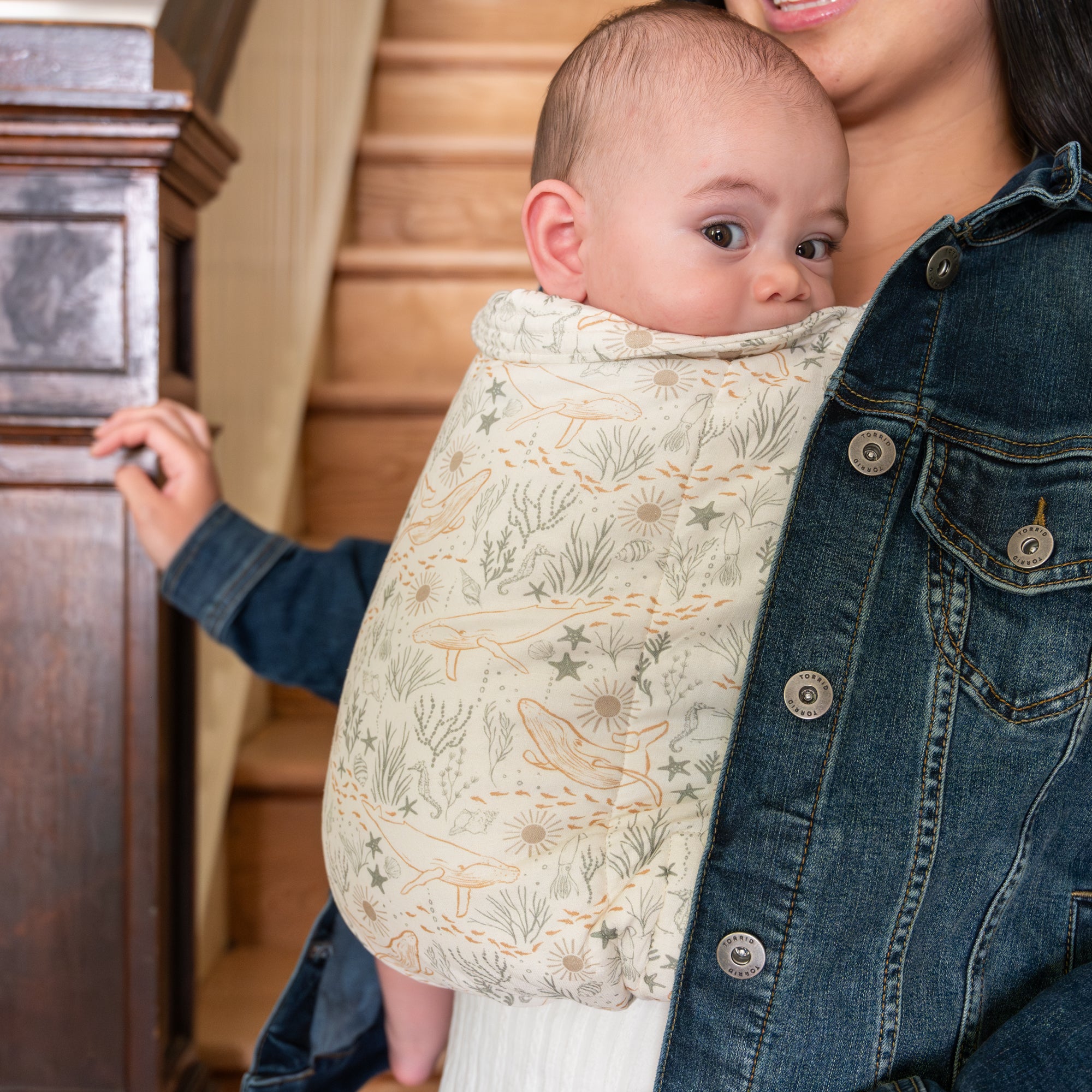 Baby in a ocean-themed baby carrier held by a person in a denim jacket.