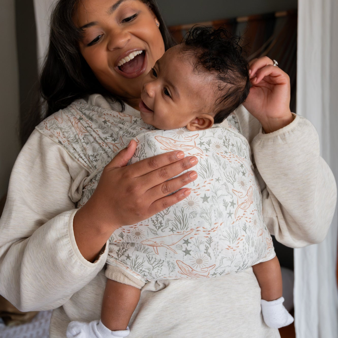Woman holding a baby in a ocean print wrap, both smiling indoors.
