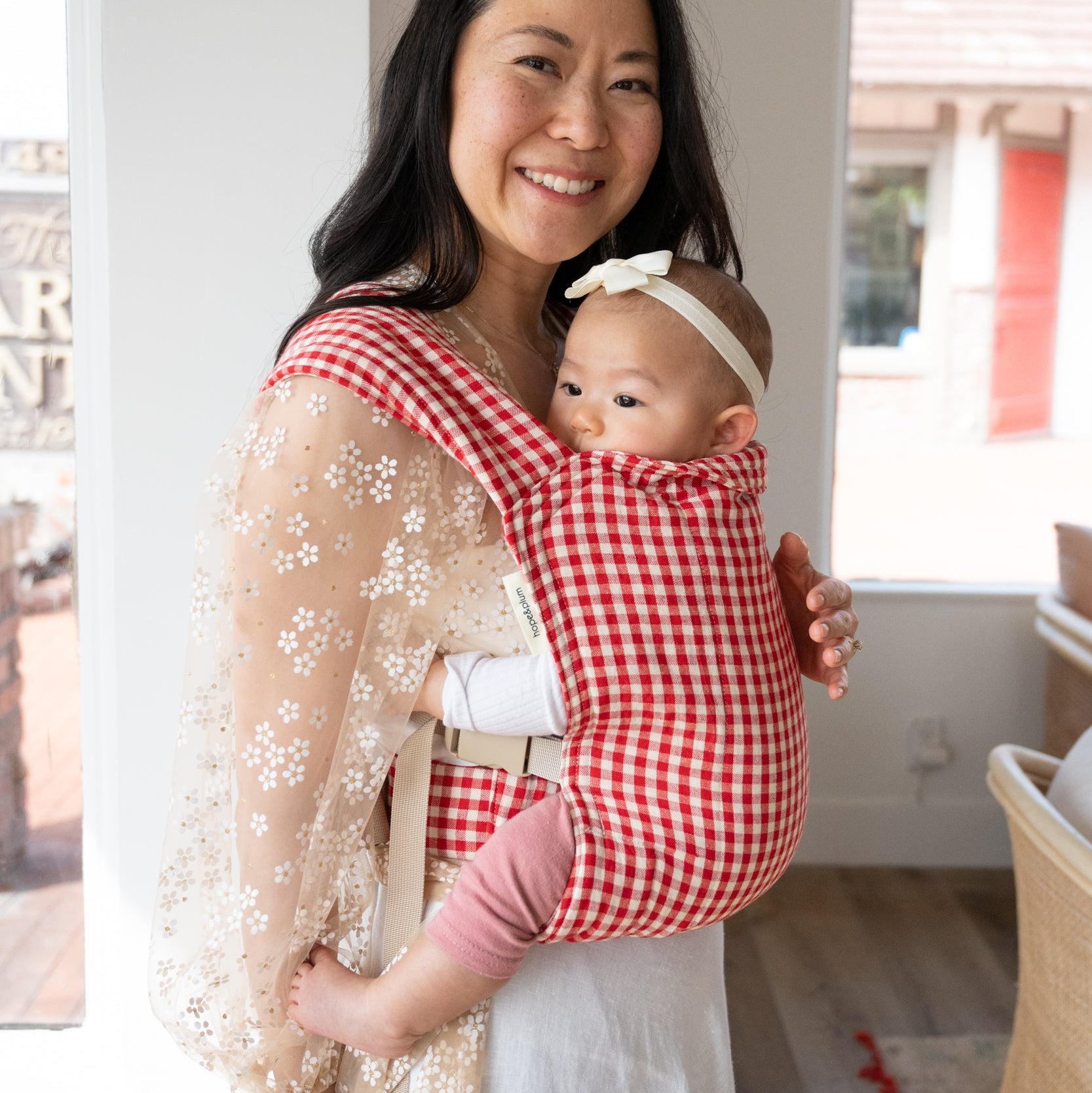 Woman holding a baby in a red and white gingham carrier indoors.