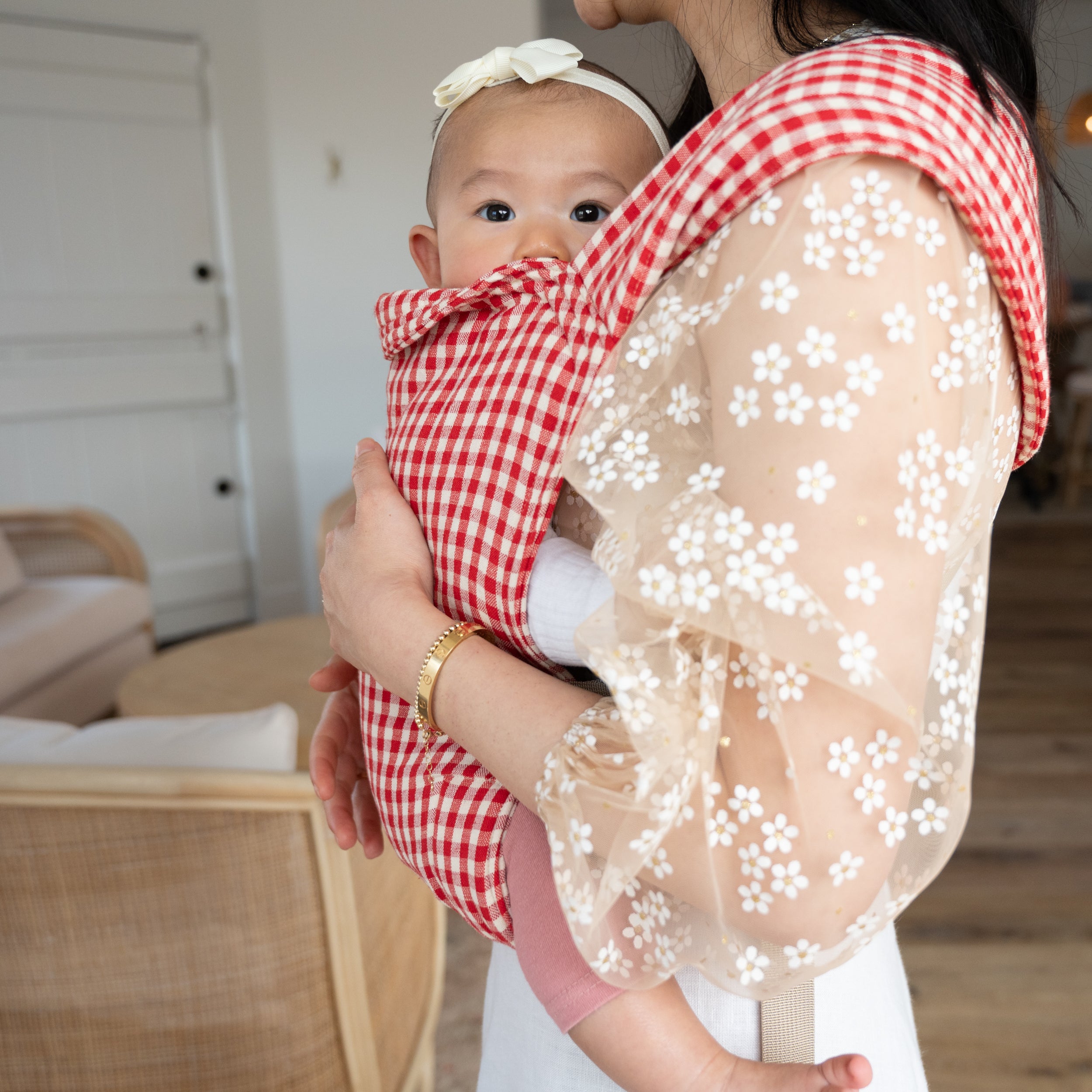 Woman holding a baby in a red and white gingham buckle carrier with a floral-patterned shirt.