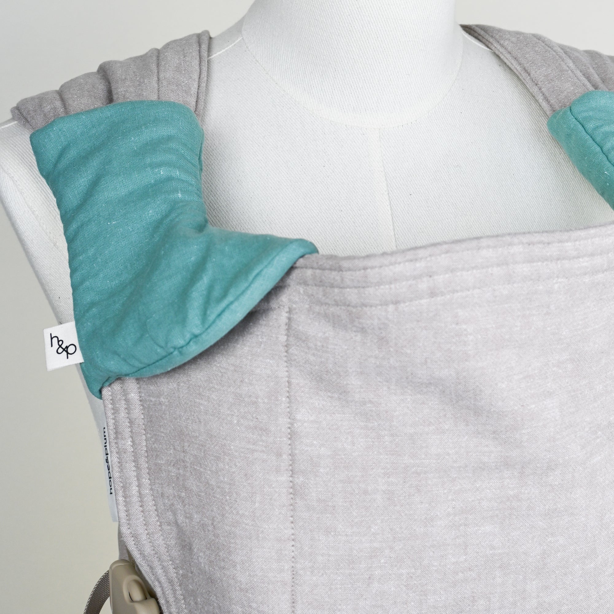 greige baby carrier with green drool pads on a mannequin, featuring 'hope&plum' label.
