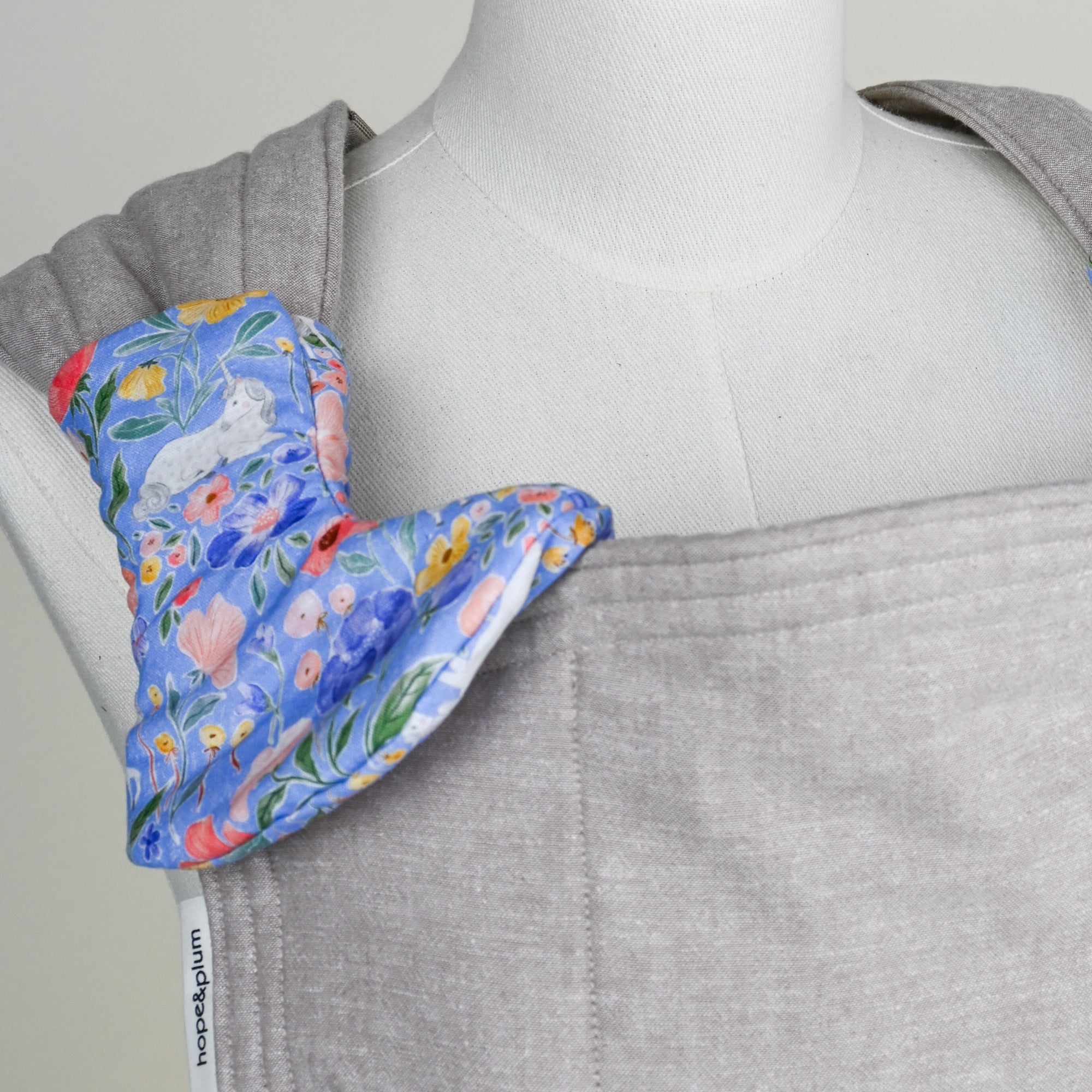 greige baby carrier with floral unicorn drool pads on a mannequin, featuring 'hope&plum' label.