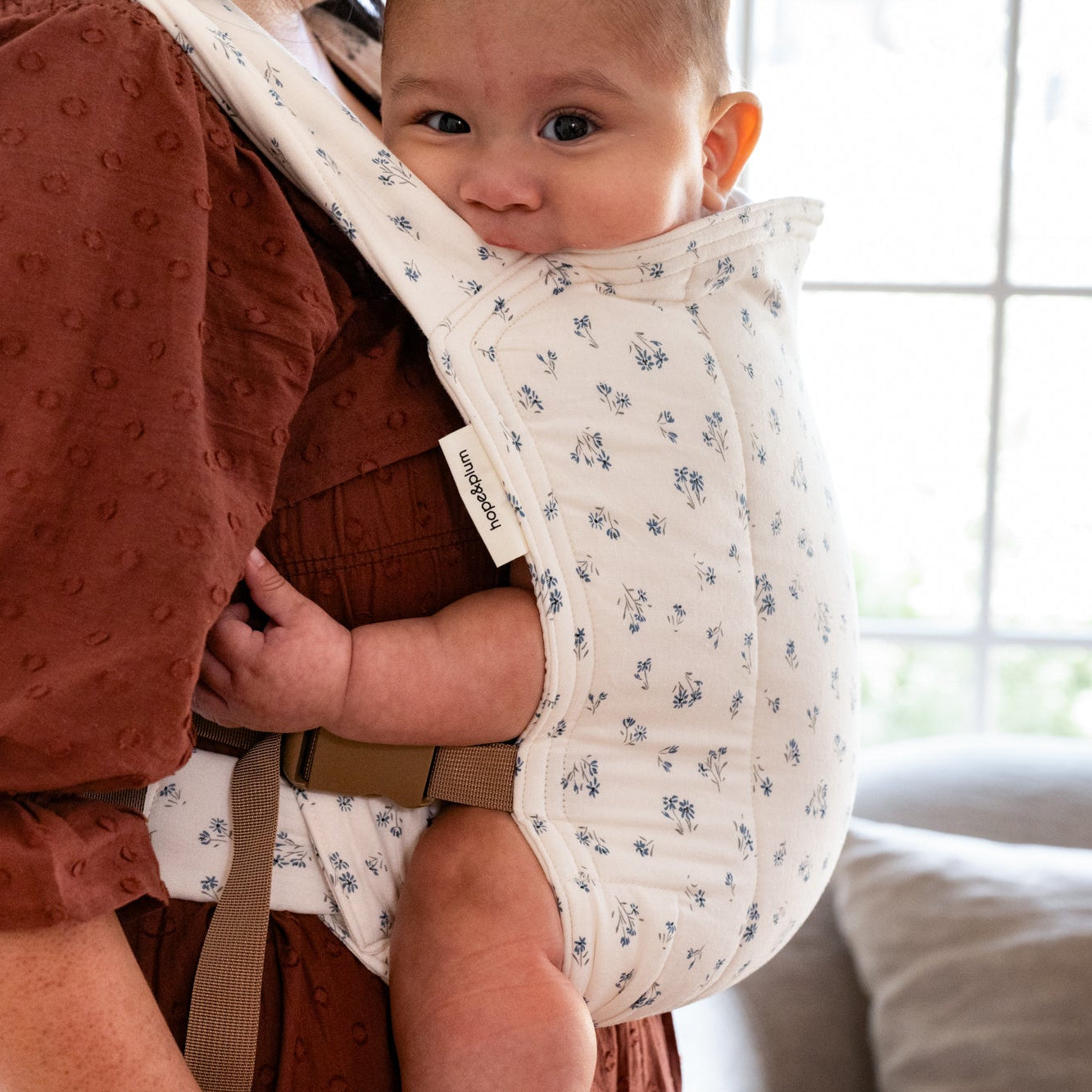 Person holding a baby in a floral baby carrier indoors.