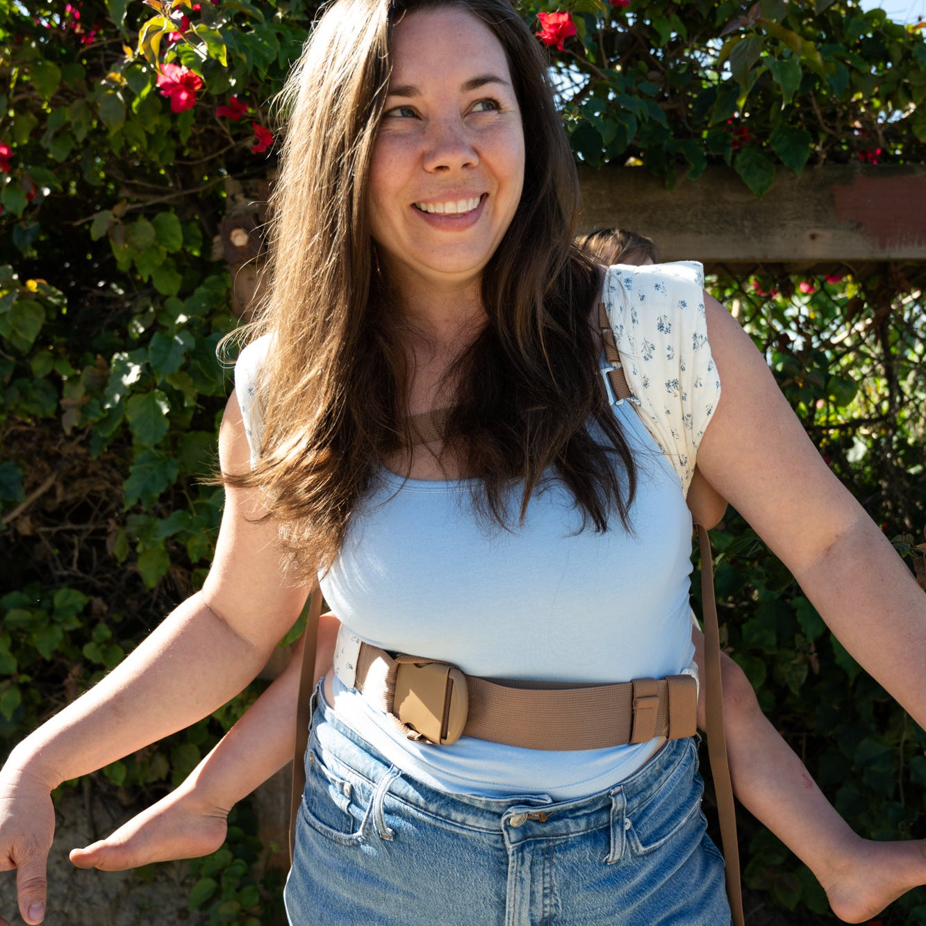 Woman standing outdoors in a light floral baby carrier with a floral arch and greenery in the background