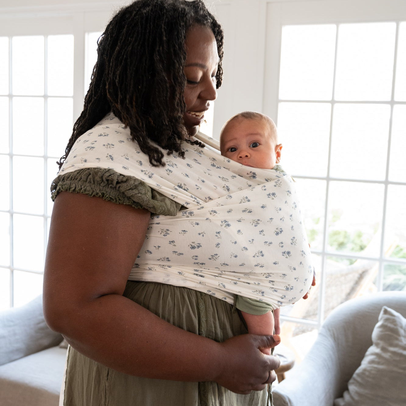 Woman holding a baby in a dainty floral wrap in a bright room with large windows.