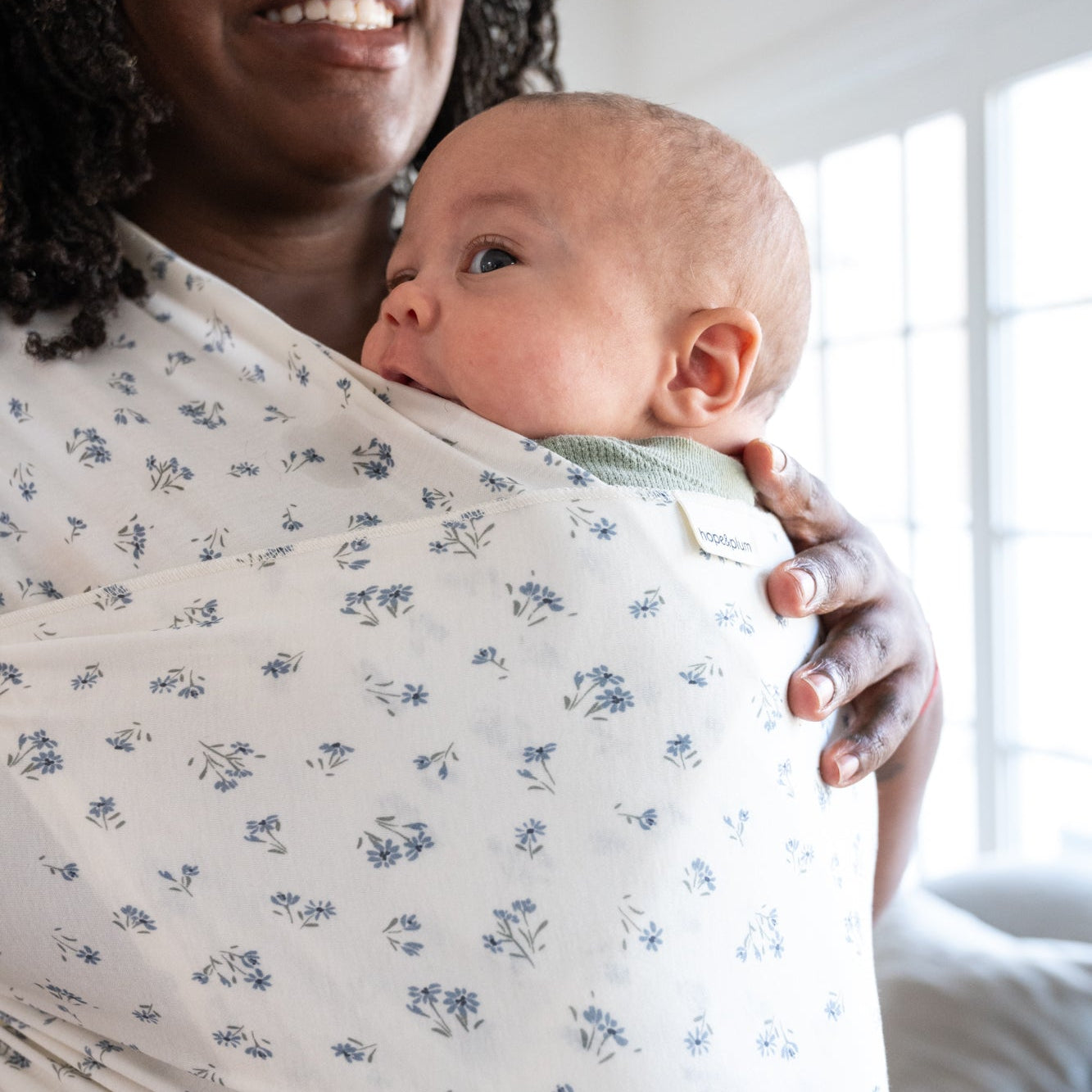 Woman holding a baby in a dainty floral wrap in a bright room with large windows.