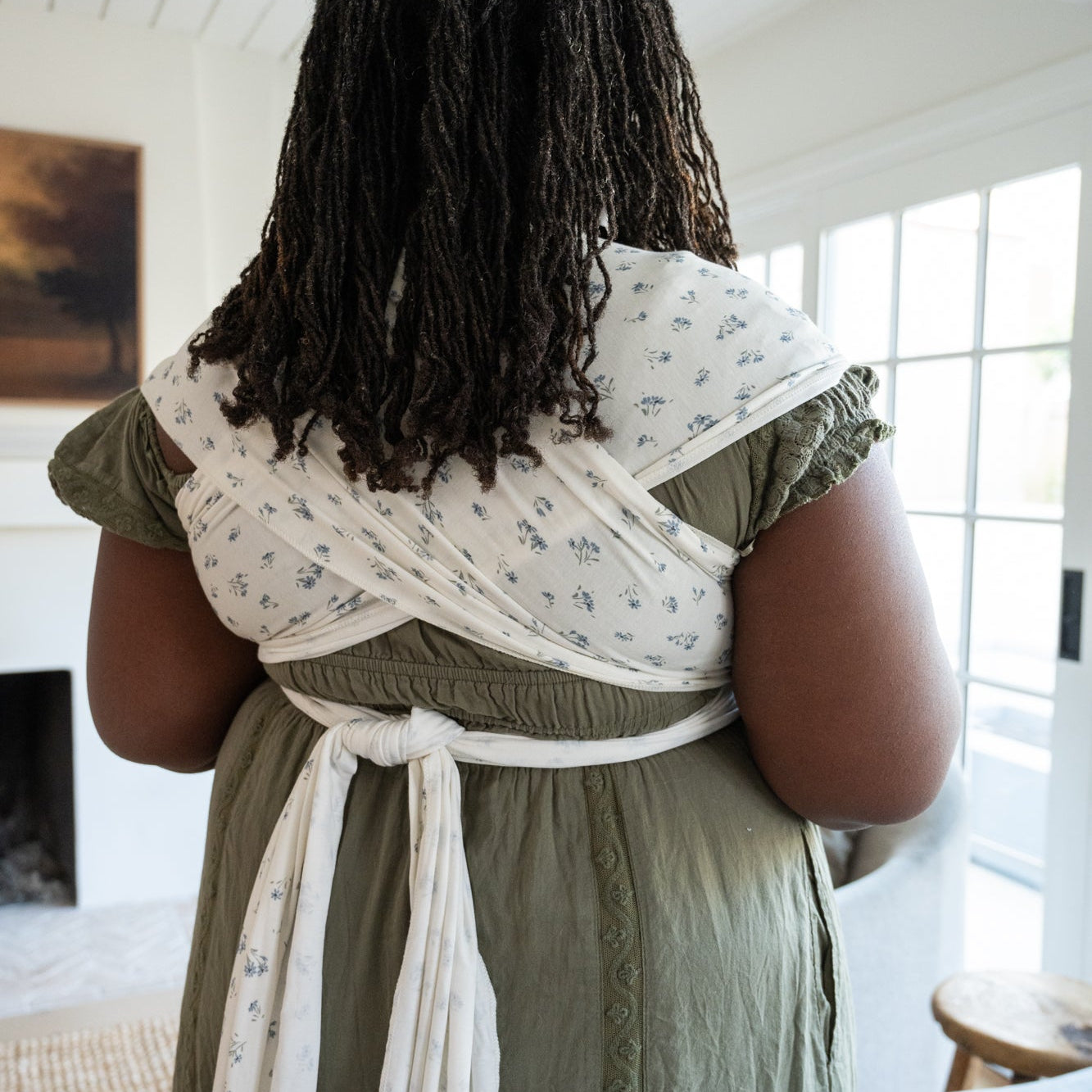 Woman holding a baby in a dainty floral wrap in a bright room with large windows.