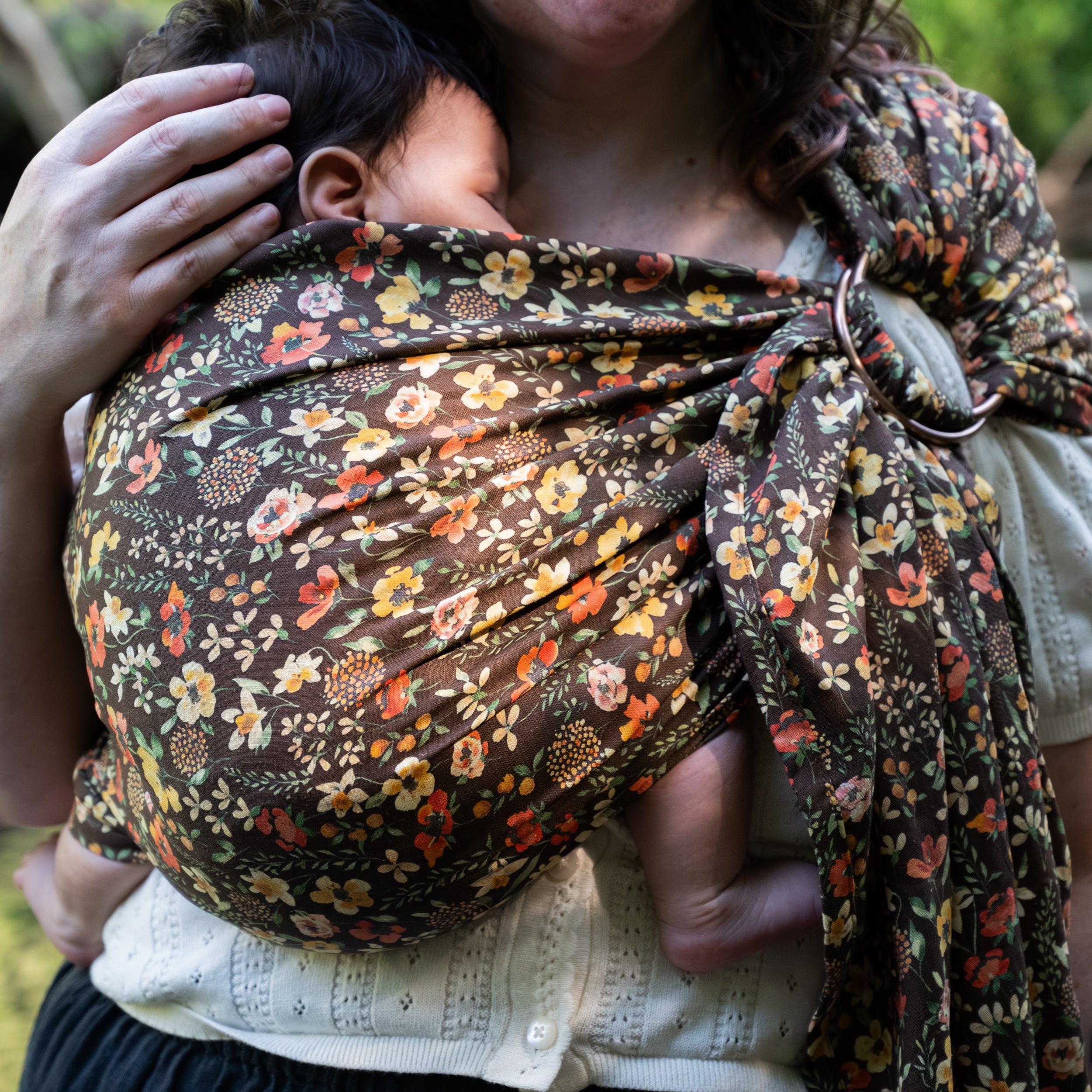 Close-up of a sleeping baby wrapped in the Ember Ring Sling, a rich, chocolate brown hemp-cotton fabric printed with autumnal watercolor florals, with the shoulder ring visible.