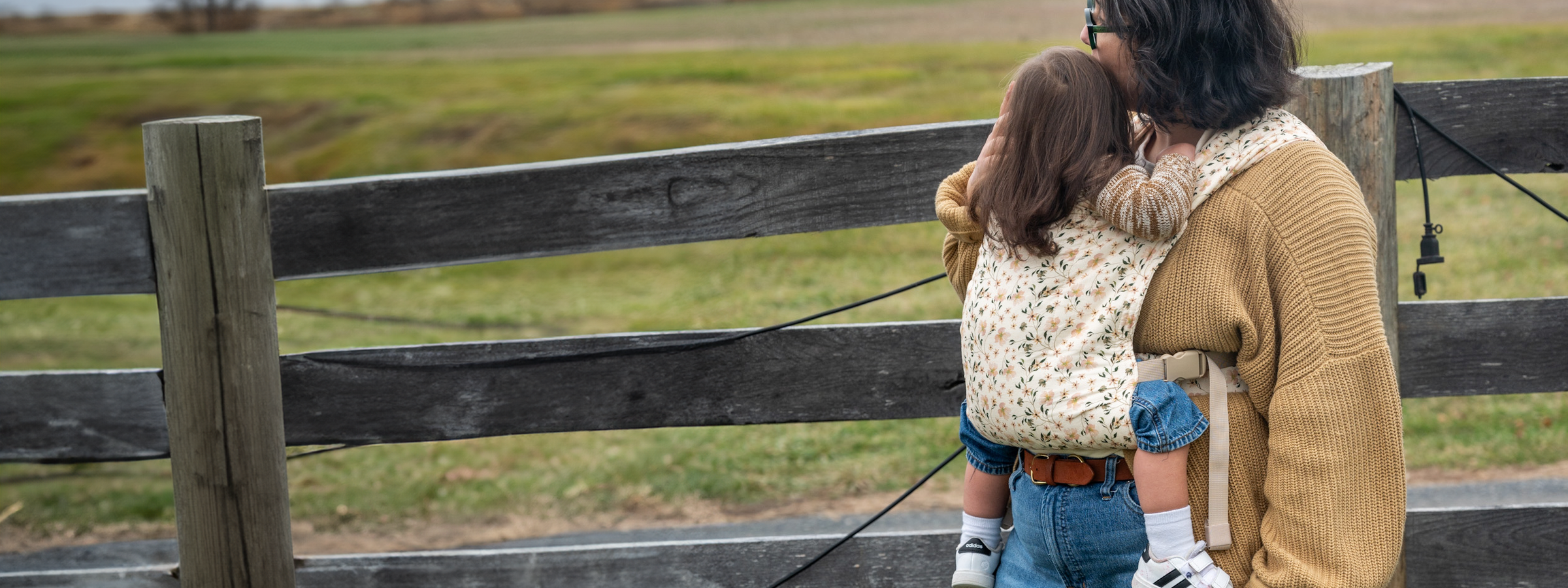 A person in a mustard yellow cardigan carries a toddler on their back in the Flor carrier — cream woven fabric printed with delicate cherry blossoms and green leaves — standing by a wooden fence overlooking a field.