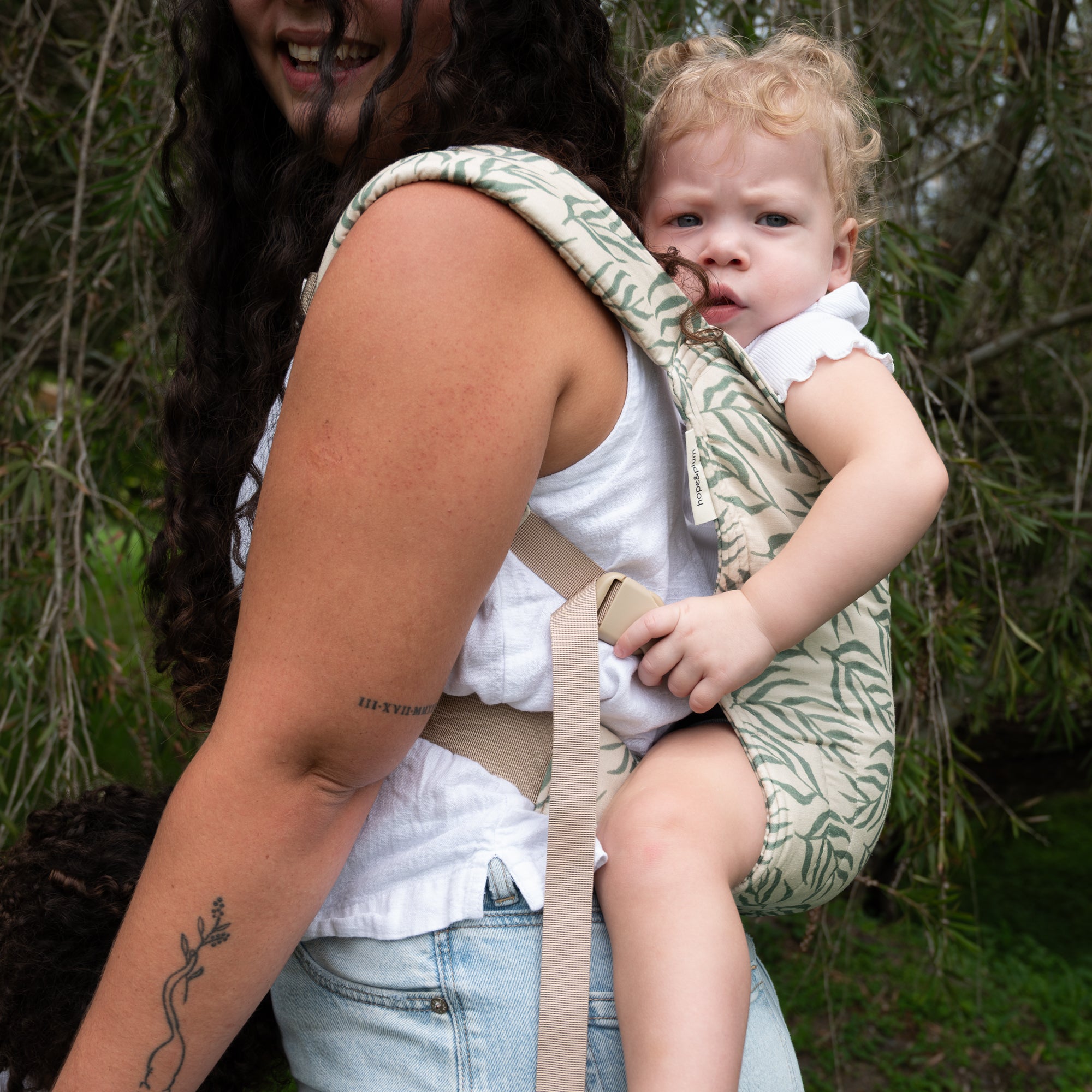 Woman carrying a baby in a leaf print baby carrier outdoors with greenery in the background